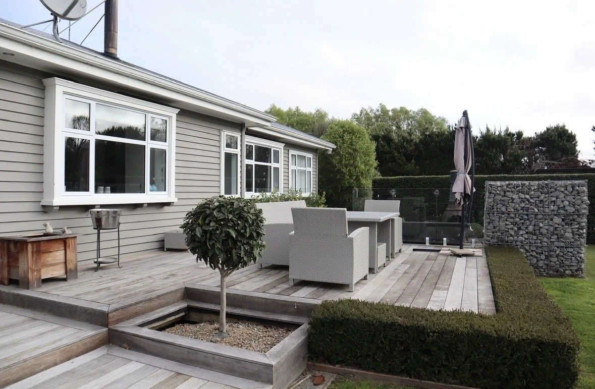 Outdoor patio with a wooden deck, white outdoor furniture, a closed umbrella, a small tree in a planter, and a stone wall privacy screen, with a house with gray siding and windows in the background.