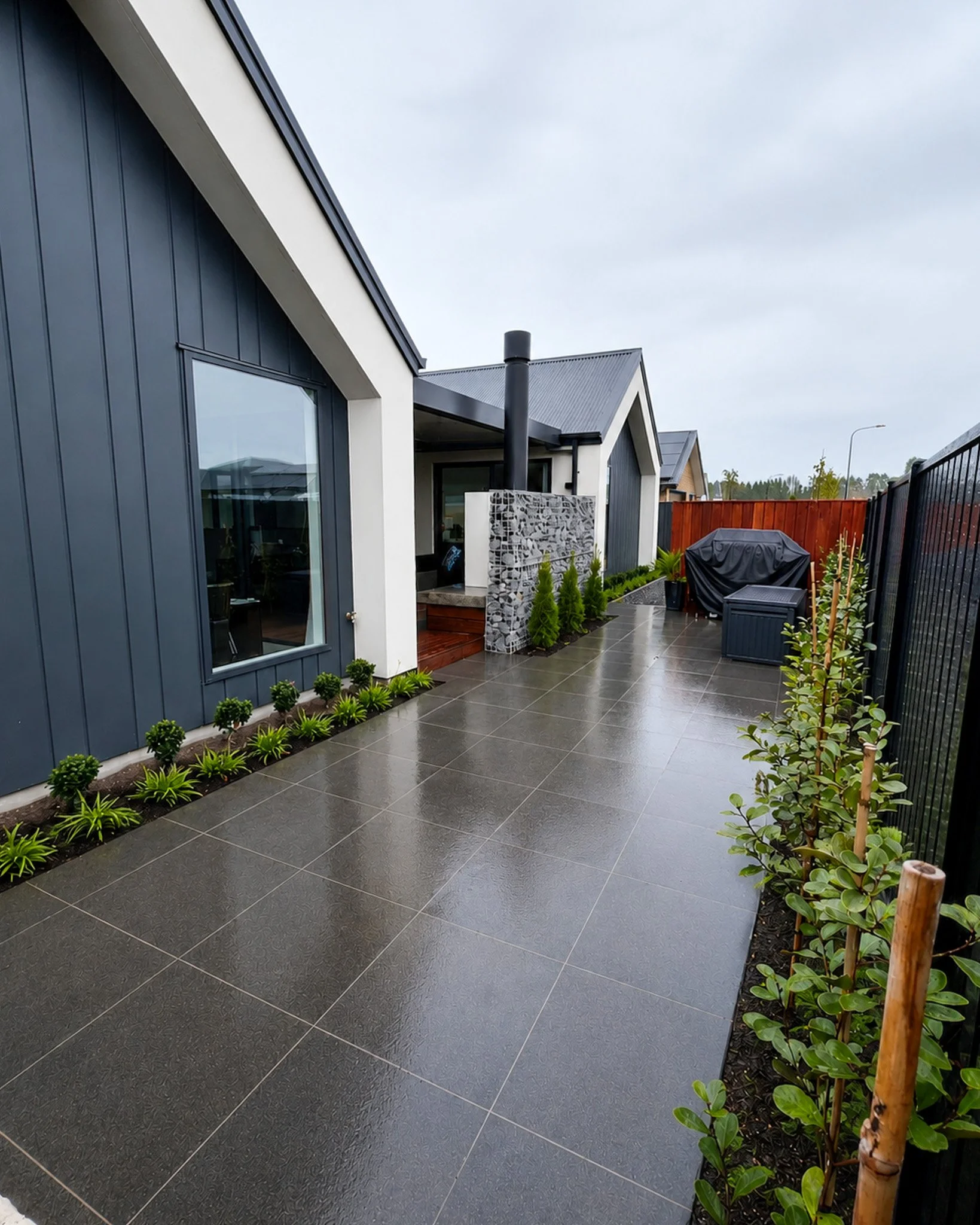 View of a modern backyard terrace with dark gray tiled flooring, bordered by small green plants, adjacent to a blue house with large glass windows, and a covered outdoor seating area, enclosed by a black metal fence, in a suburban neighborhood with o