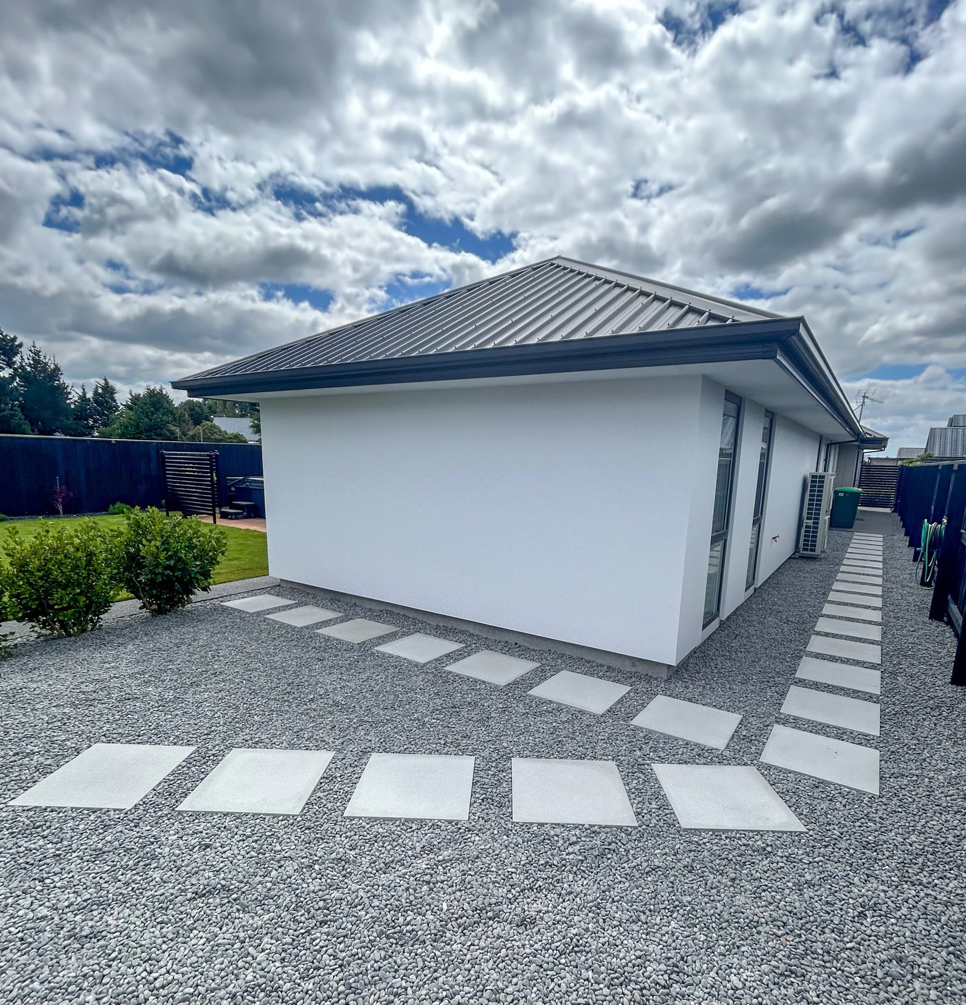 Modern house with a metal roof, white exterior walls, large windows, surrounded by a gravel yard, with a pathway of square concrete tiles, green bushes, trees, and a cloudy sky overhead.
