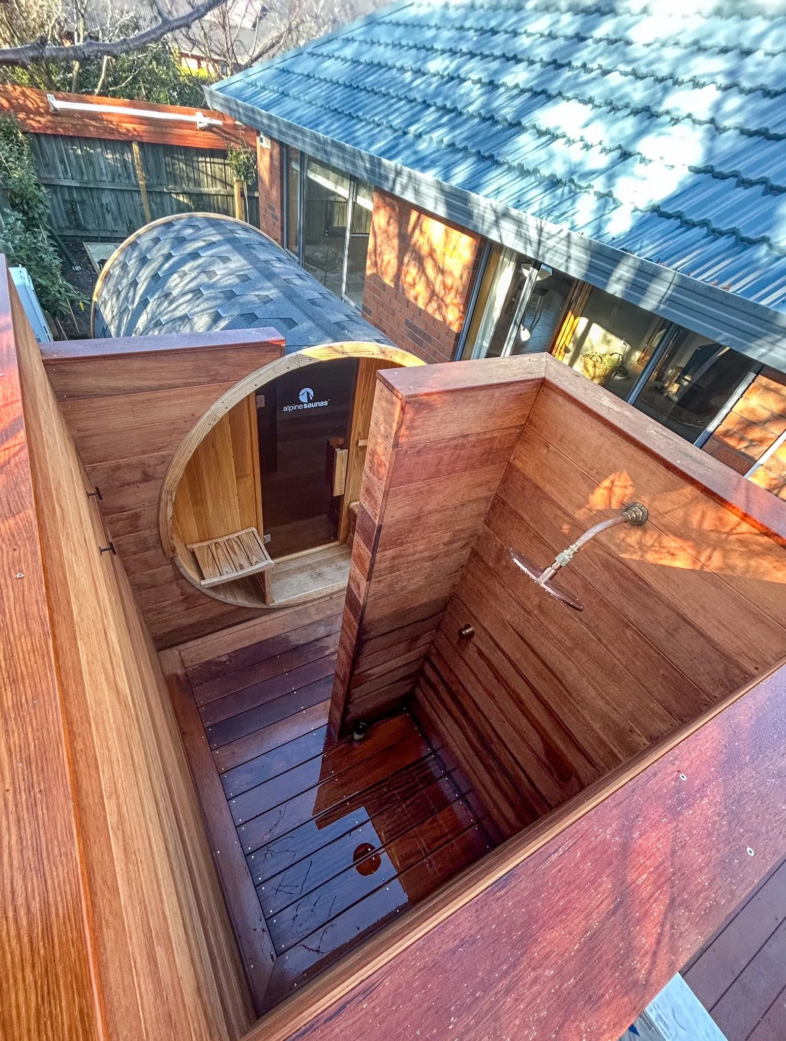 View from above of a backyard with a wooden hot tub, a small wooden sauna with an arched door, a shed with a sloped metal roof, and a house with large windows. A wooden fence surrounds the yard, and there are trees casting shadows.