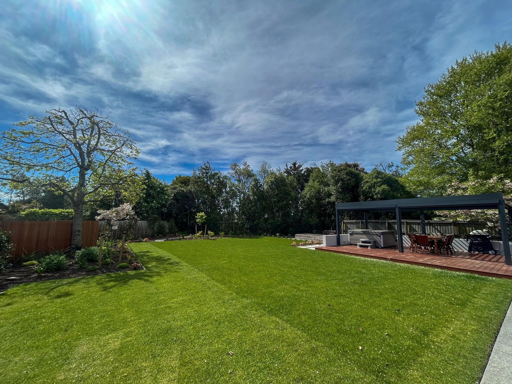 A backyard with a lush green lawn, a flowering tree, trees along the fence line, a covered patio area with outdoor furniture, and a partly cloudy blue sky with sunlight