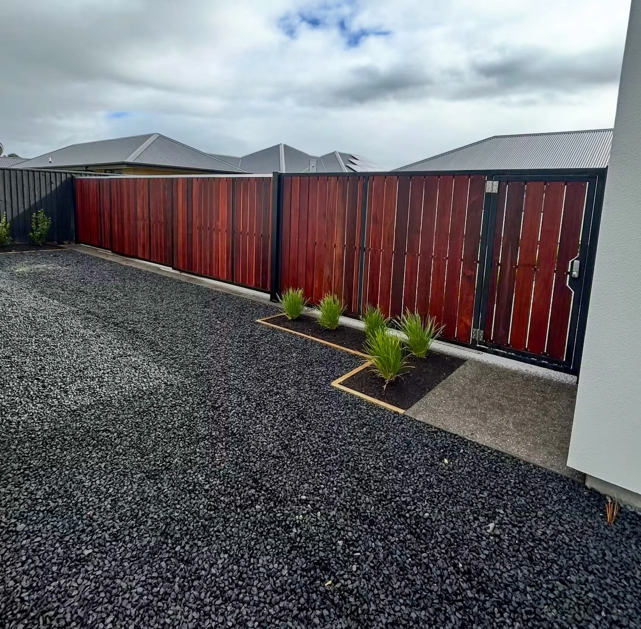 View of a backyard with a tall red wooden fence, small green plants, gravel ground, and rooflines of neighboring houses under a cloudy sky.