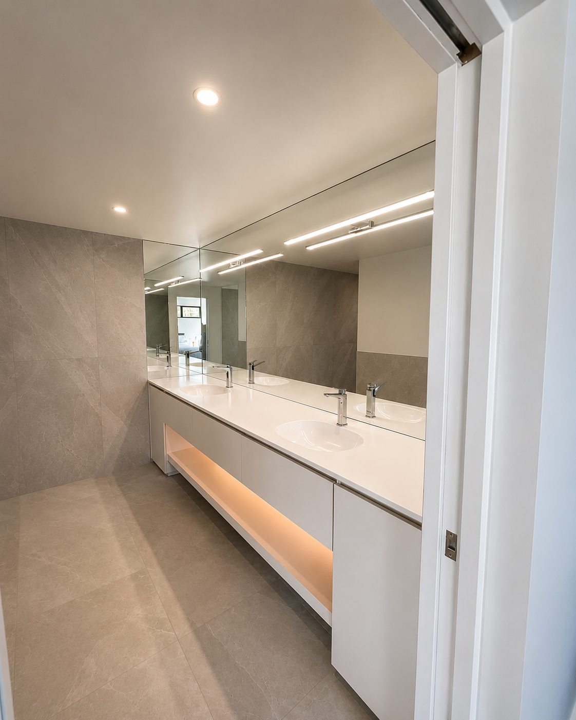 Modern bathroom with a long white vanity, four sinks, large mirror, beige tile walls and flooring, ceiling lights, and a door slightly open.