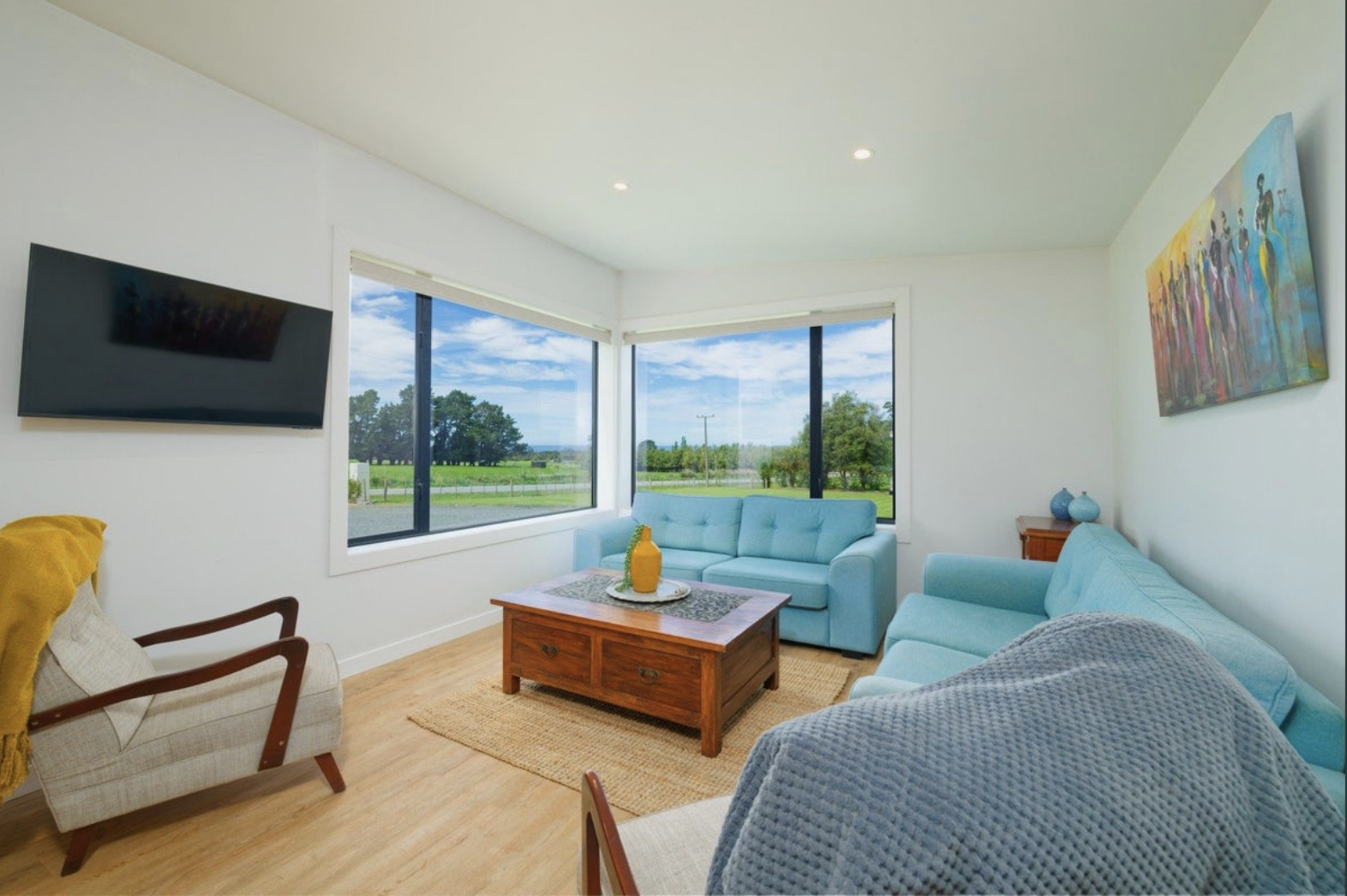 Bright living room with large windows showing a green outdoor landscape, light blue sofas, a wooden coffee table, a wall-mounted TV, and colorful artwork on the wall.
