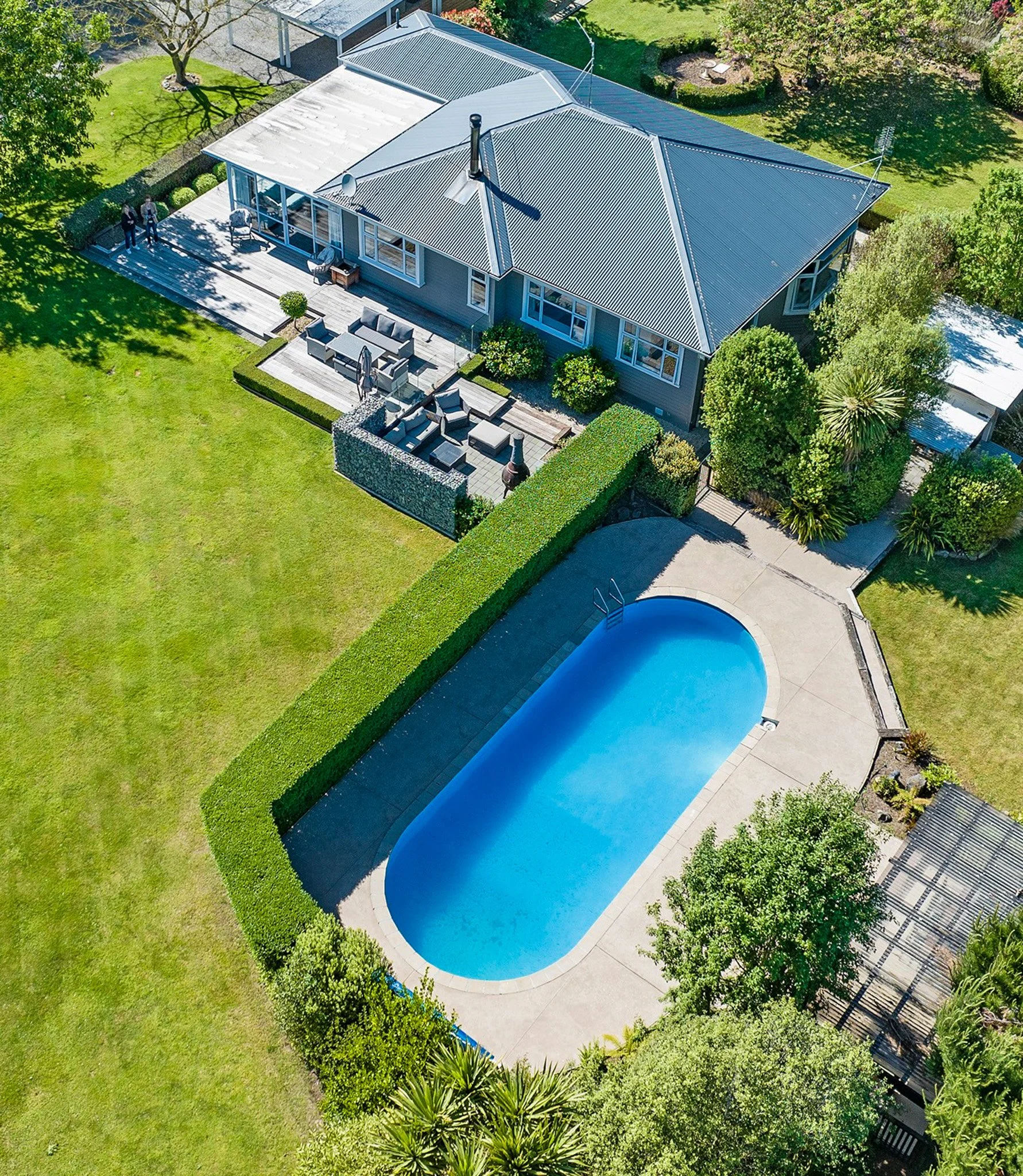 Aerial view of a house with a metal roof, a large backyard with a swimming pool, a deck with outdoor furniture, lush green lawn, trees, and landscaping.