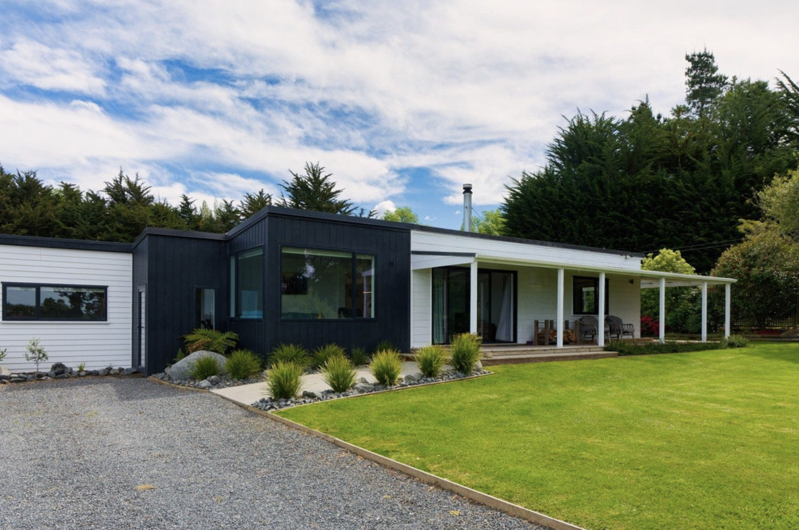 Modern house with a black and white exterior, large windows, green lawn, and a covered porch with outdoor seating, surrounded by trees.