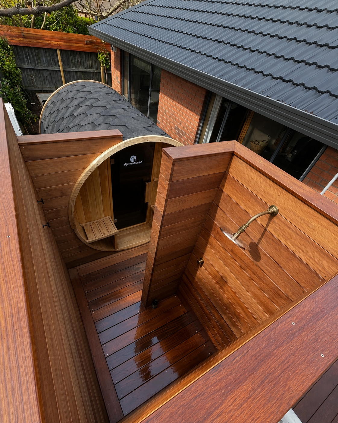 An outdoor wooden shower with a brass showerhead, enclosed by wooden walls, next to a building with brick walls and a gray metal roof, with a curved outdoor sauna structure in the background.