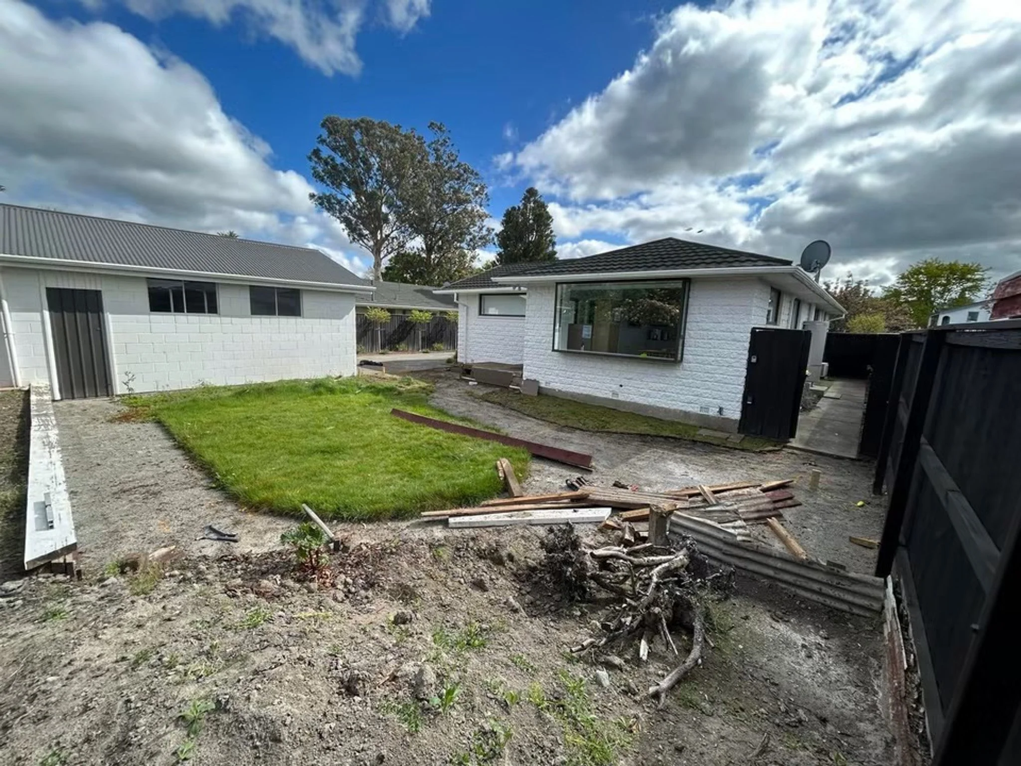 A backyard with a patch of grassy lawn, surrounded by a white brick house on the right with large windows, and a white wall with a gray metal roof on the left. The yard has patches of dirt and construction debris, indicating ongoing work. The sky is 