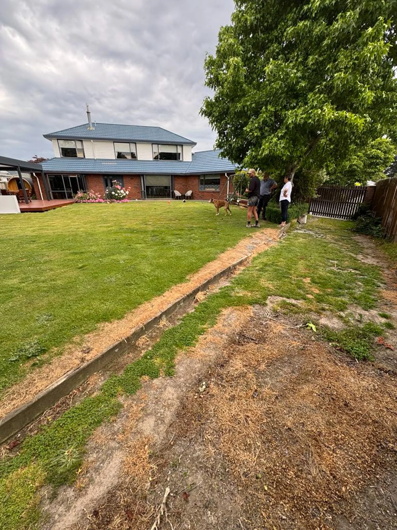 Backyard with lawn and garden bed, people standing by a tree, house in background, cloudy sky.