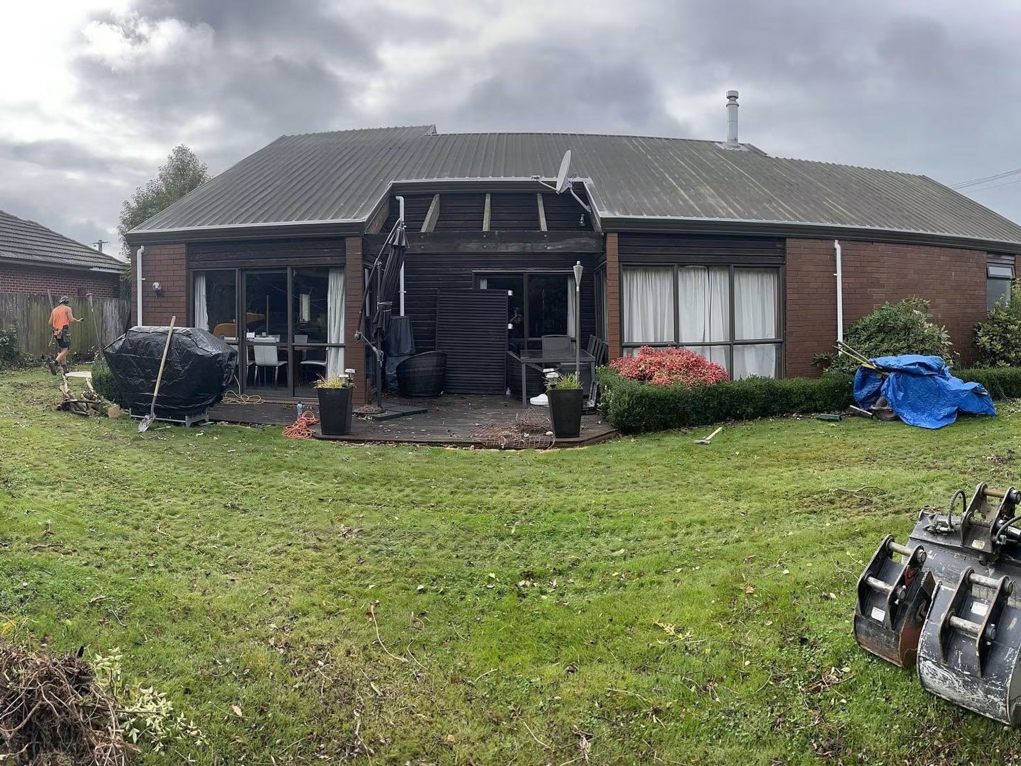 The image depicts the back view of a two-story house with a metal roof, some of which is under repair or replacement. The house has large windows with curtains. In the backyard, there is a patio with outdoor furniture, and tools and equipment scatter