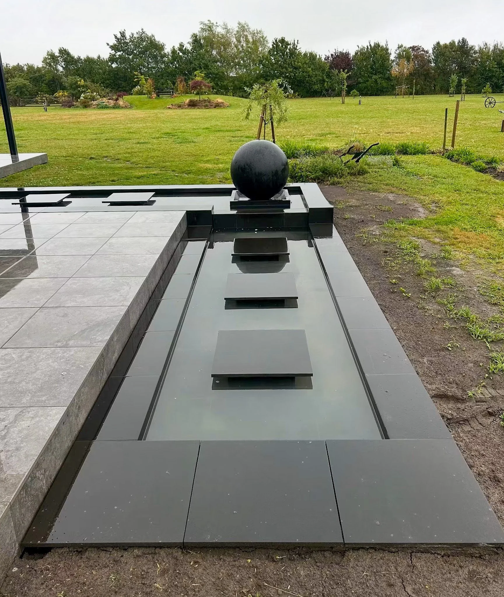 A large black globe sculpture is placed on a black tiled table outdoors, with a garden and trees in the background.