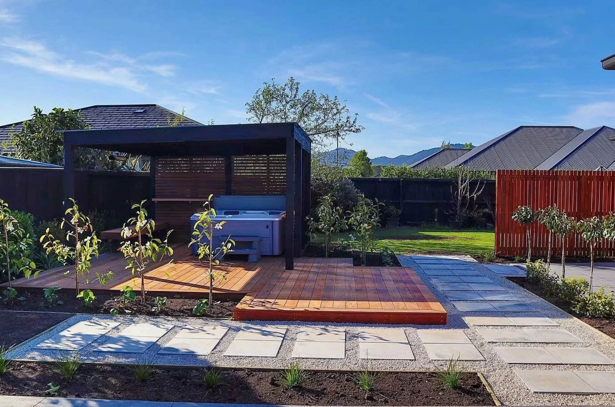 Backyard with wooden deck, pergola, plants, and mountain view under blue sky.