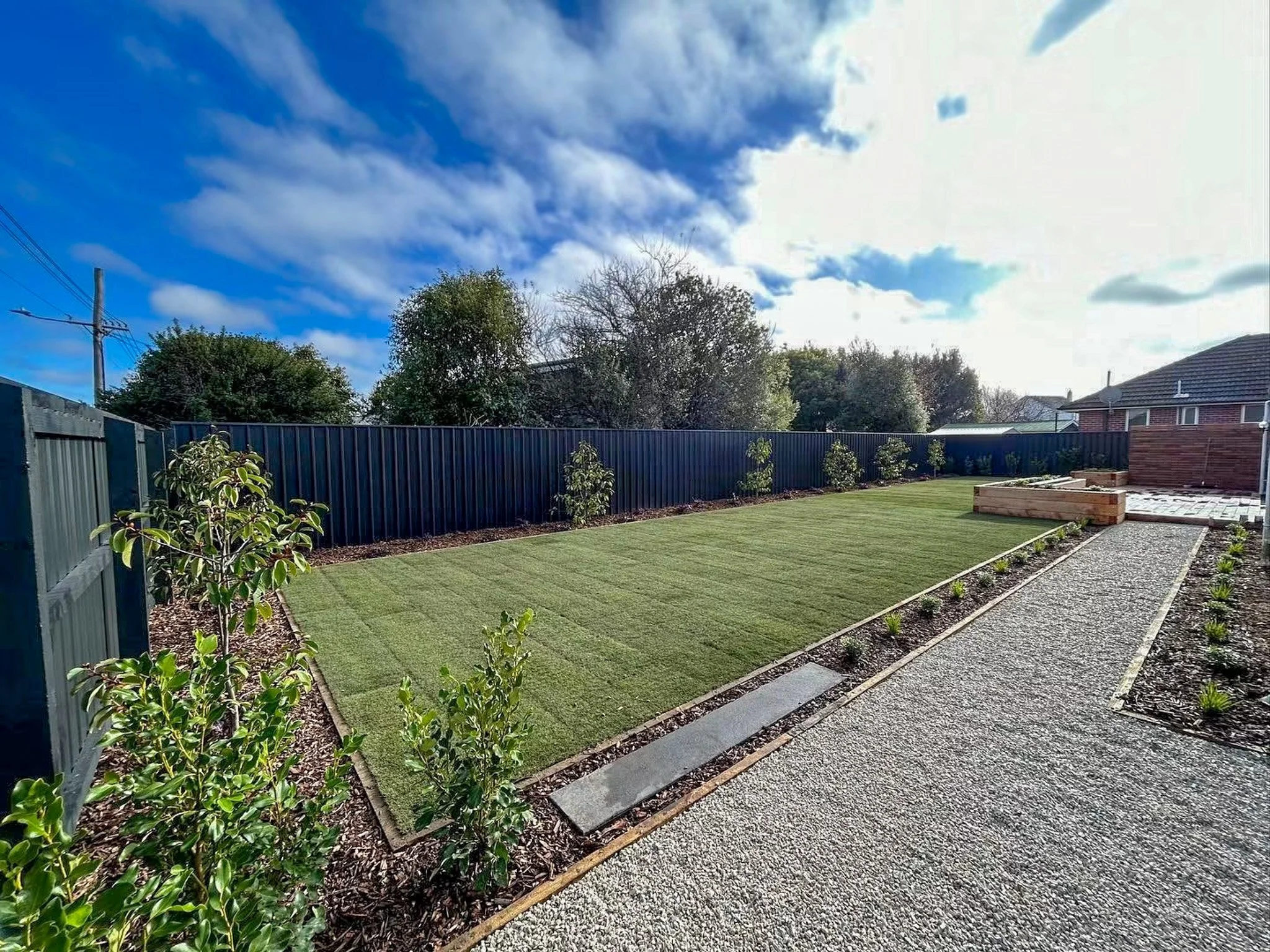A backyard with a neatly mowed lawn, small plants, a pathway with stepping stones, a gravel area, trees and a metal fence, under a partly cloudy sky.