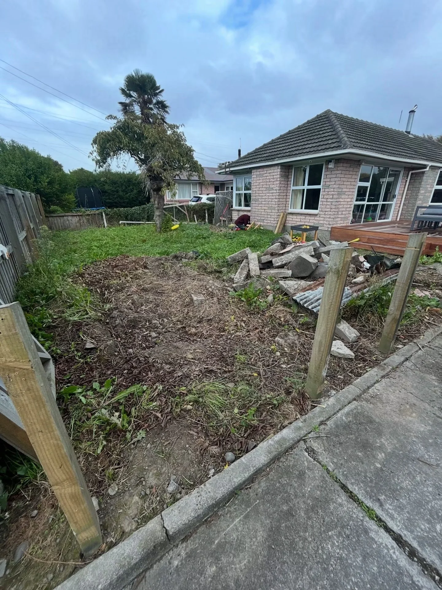 A backyard with a house, tree, and a pile of bricks, with some wood and tools, and a person working near the house.
