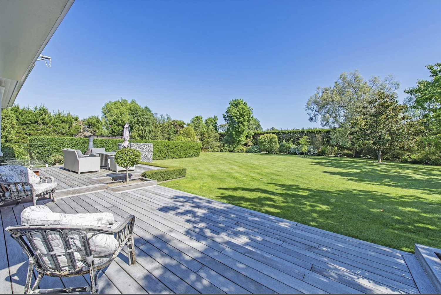 View of a backyard with a wooden deck, outdoor seating, and a large green lawn surrounded by trees and bushes under a clear blue sky.