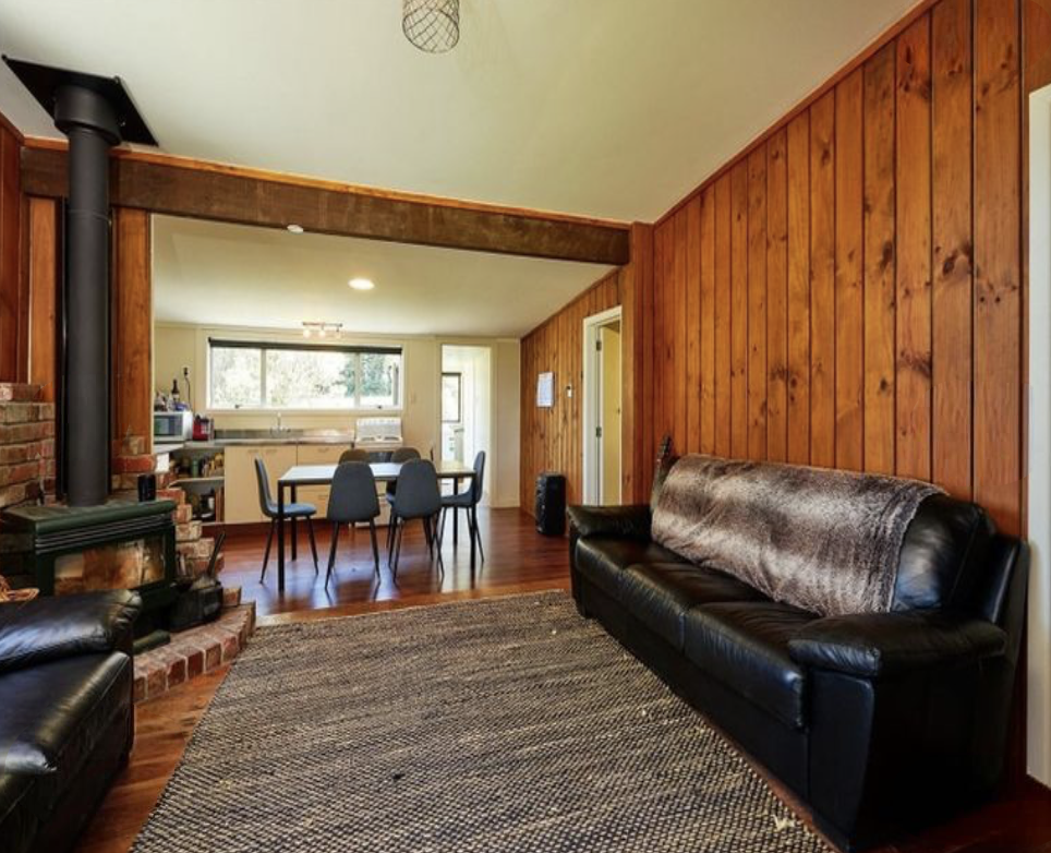 Living room with wood-paneled walls, a black leather sofa with a fur throw, a patterned area rug, and a wood fireplace with a black chimney pipe, opening into a dining area with a table and six chairs, and a kitchen in the background.