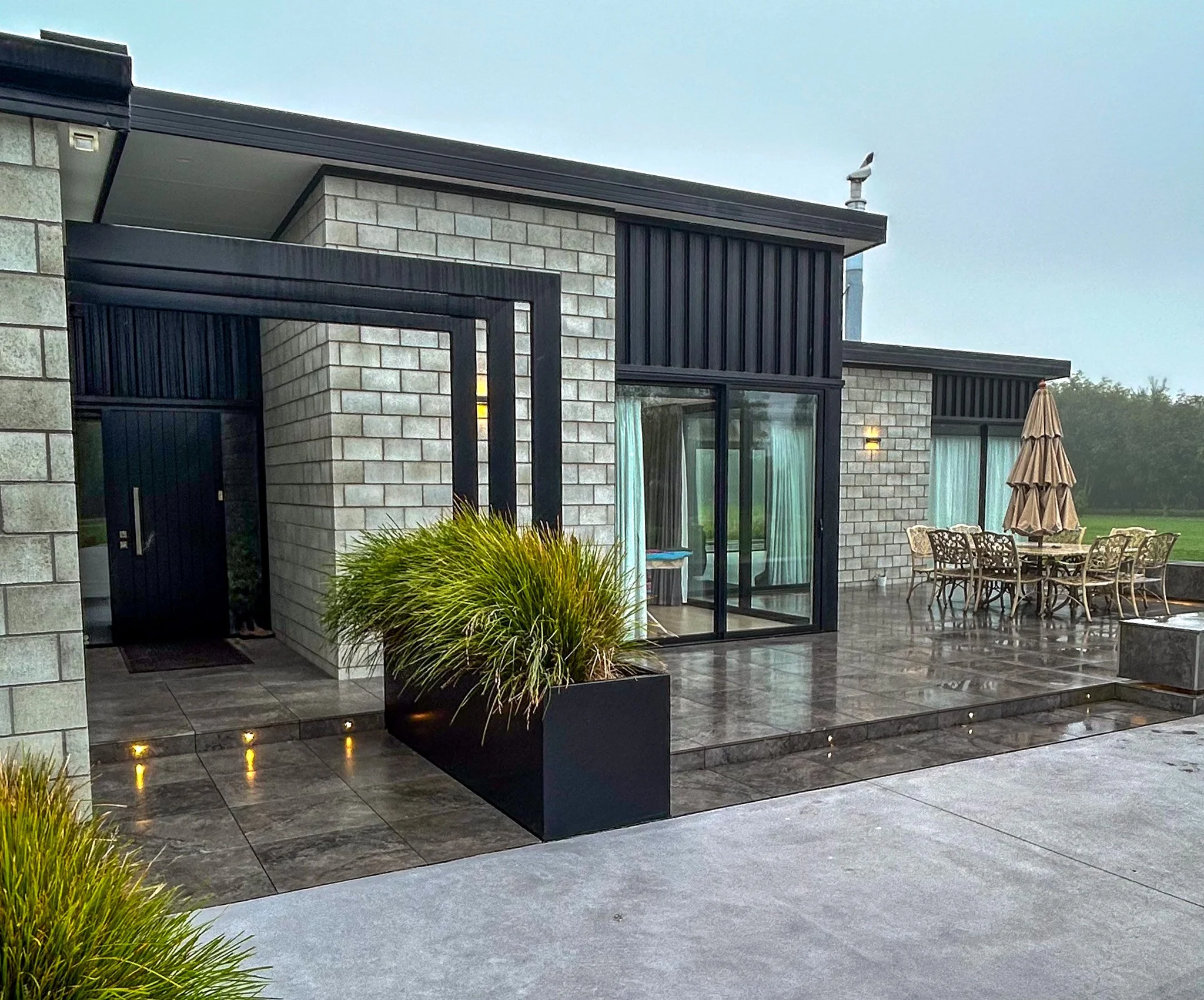 Modern house with brick and black metal siding, glass sliding doors, outdoor dining table and chairs, patio umbrella, potted plants, and pathway with small lights.