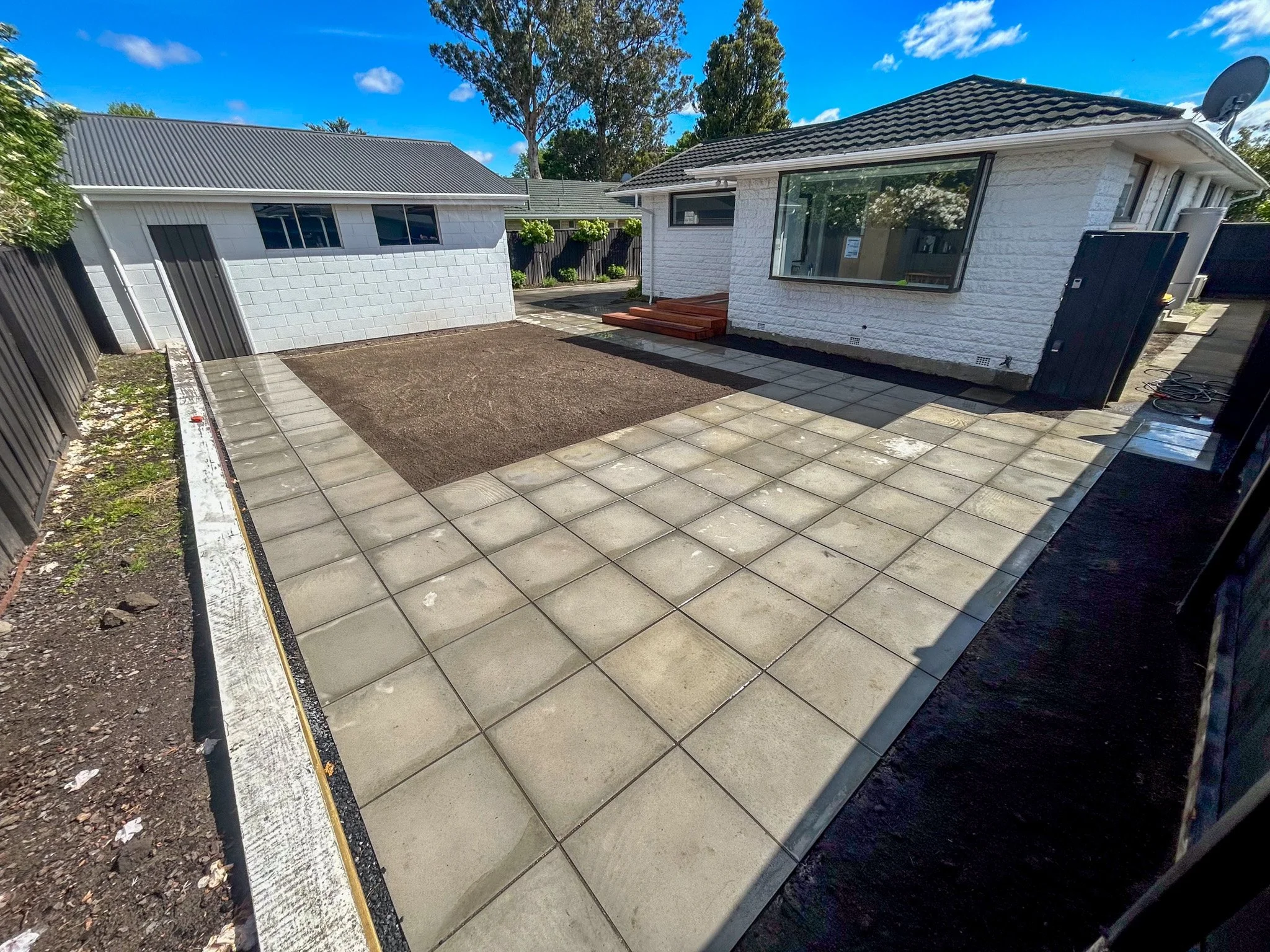 Backyard with a small garden bed, paved area, and a house with large windows and a tiled roof under a blue sky.