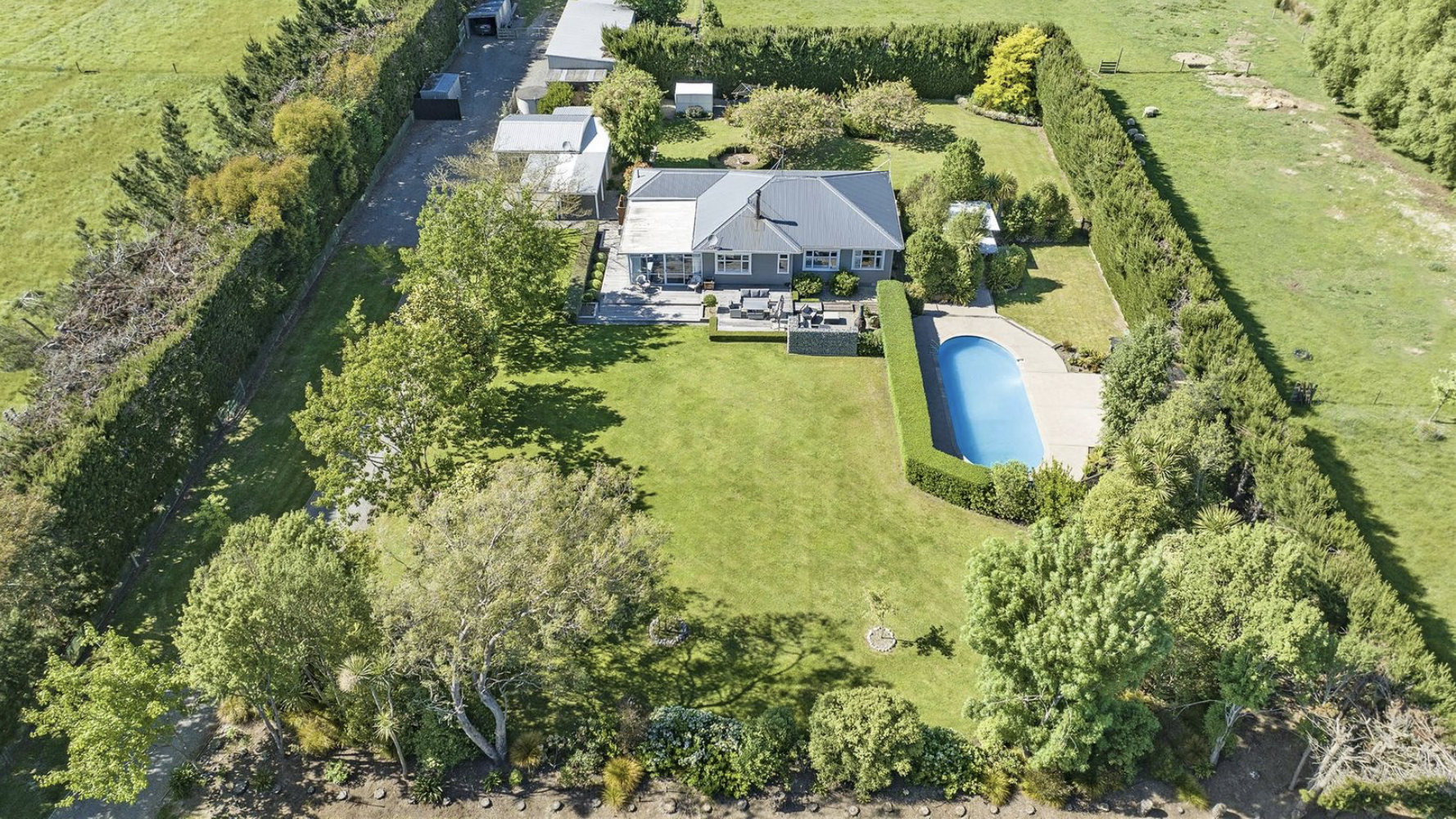 Aerial view of a backyard with a house, a swimming pool, a large lawn, trees, and a hedge fence.