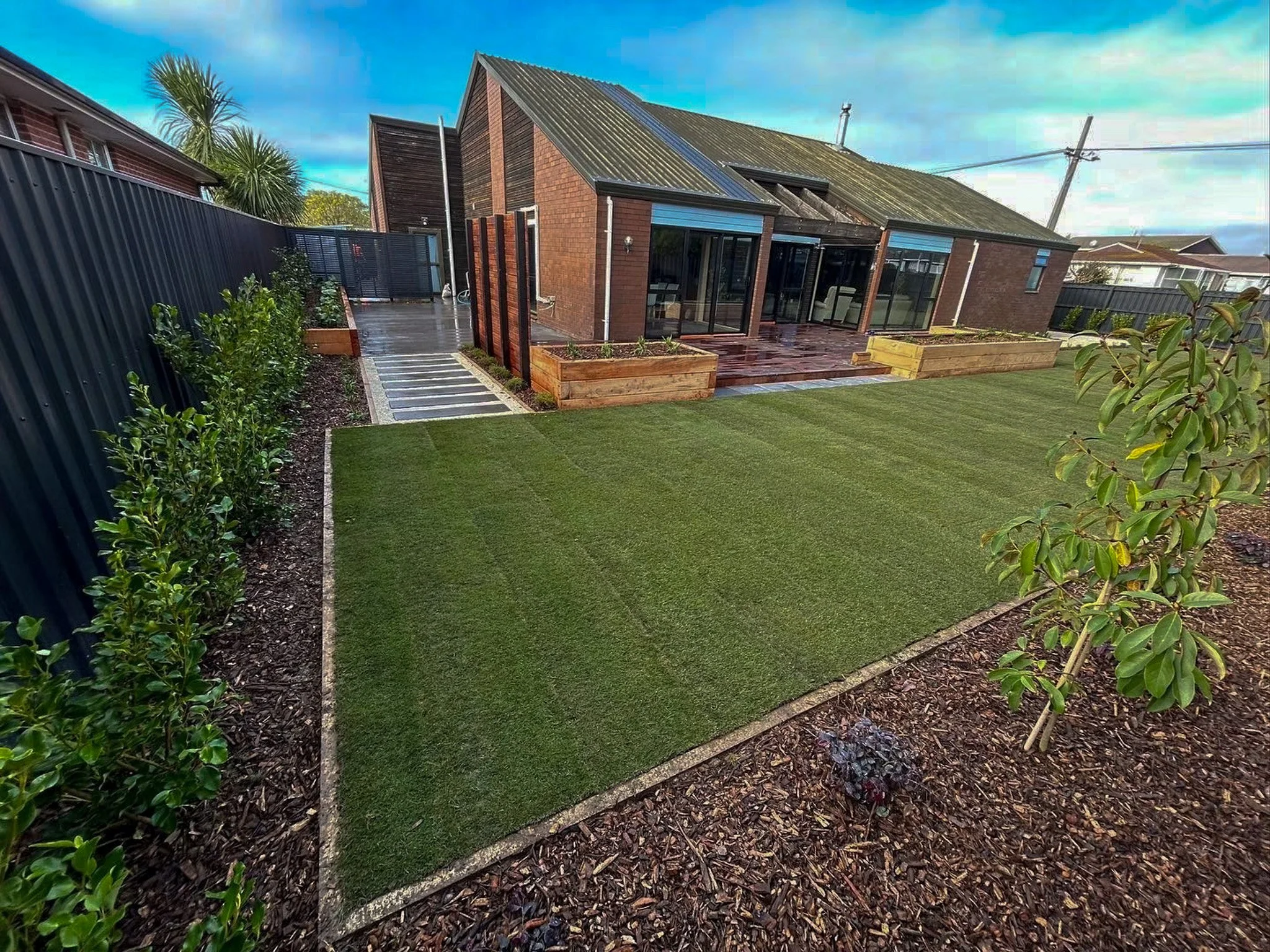 A backyard with a artificial grass lawn, elevated garden beds, and a brick house with a glass enclosure, a wooden deck, and a metal roof.