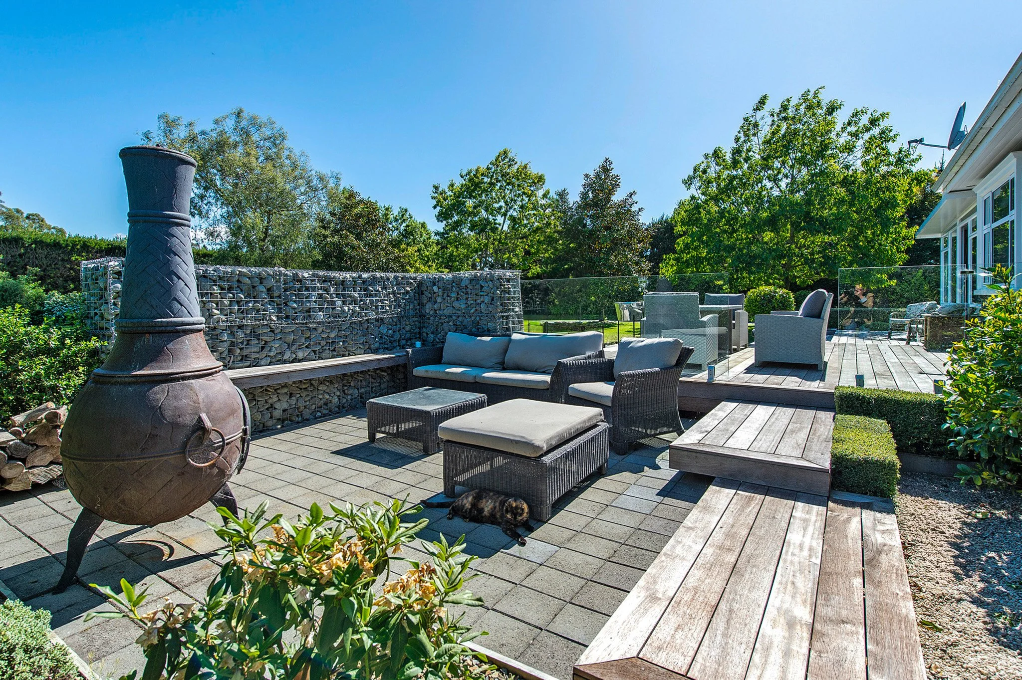 A wooden outdoor patio with patio furniture, a chiminea, a stone and wire gabion fence, lush green trees, and a clear blue sky.