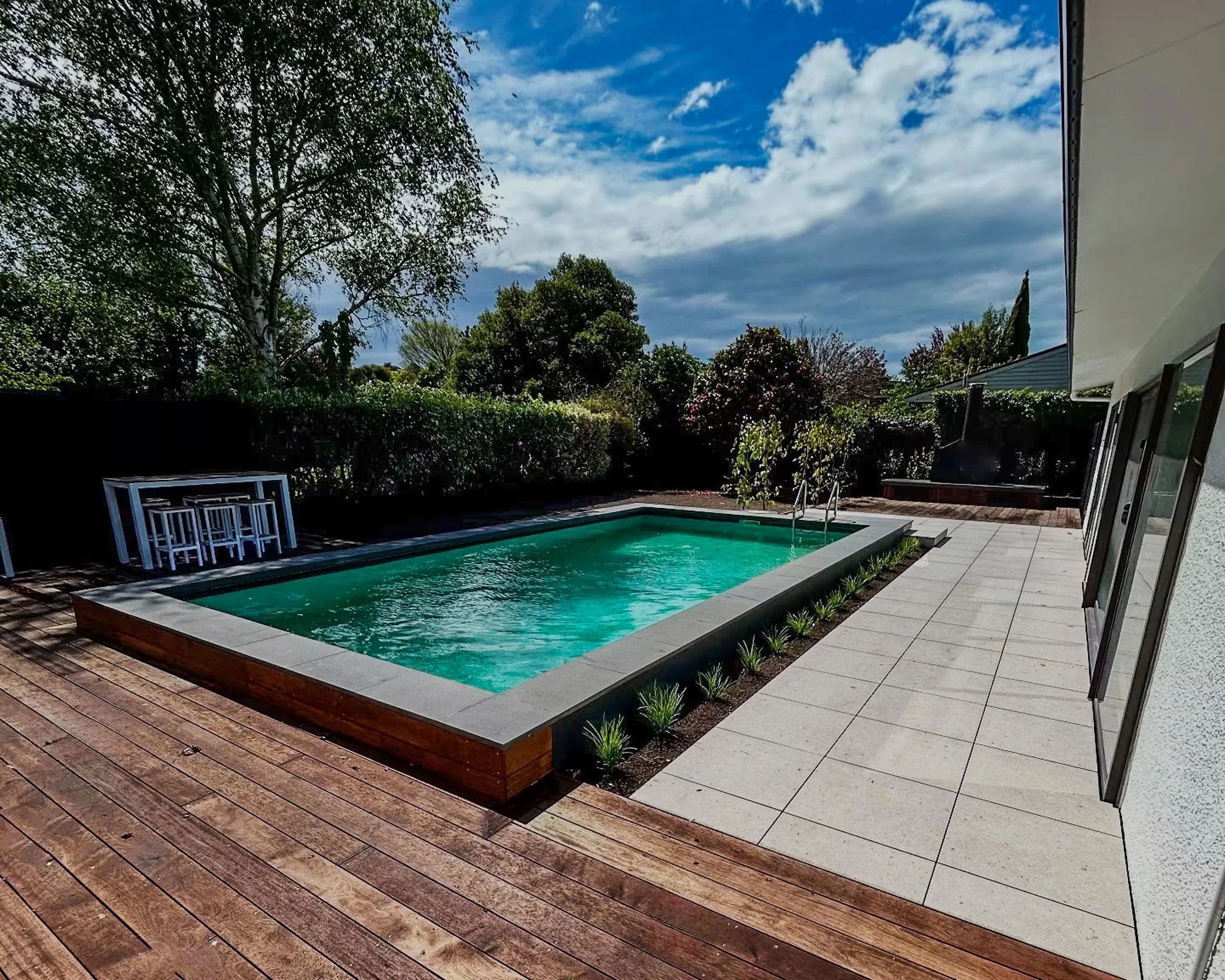 A backyard with a small swimming pool, surrounded by a wooden deck and a tiled patio. There are trees and bushes in the background with a partly cloudy sky overhead.