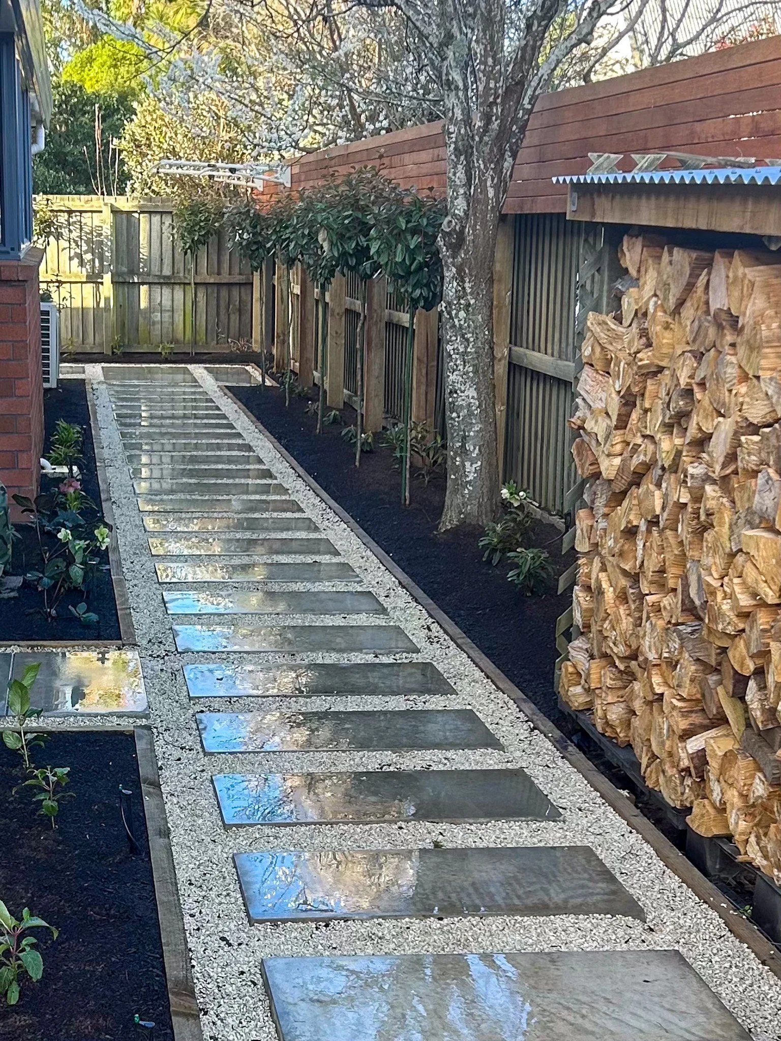 A garden pathway with rectangular stone tiles set in gravel, bordered by black soil with small plants. On the right, there's a stack of chopped firewood next to a wooden fence and a shed with a metal roof. On the left, a garden bed with plants and a 