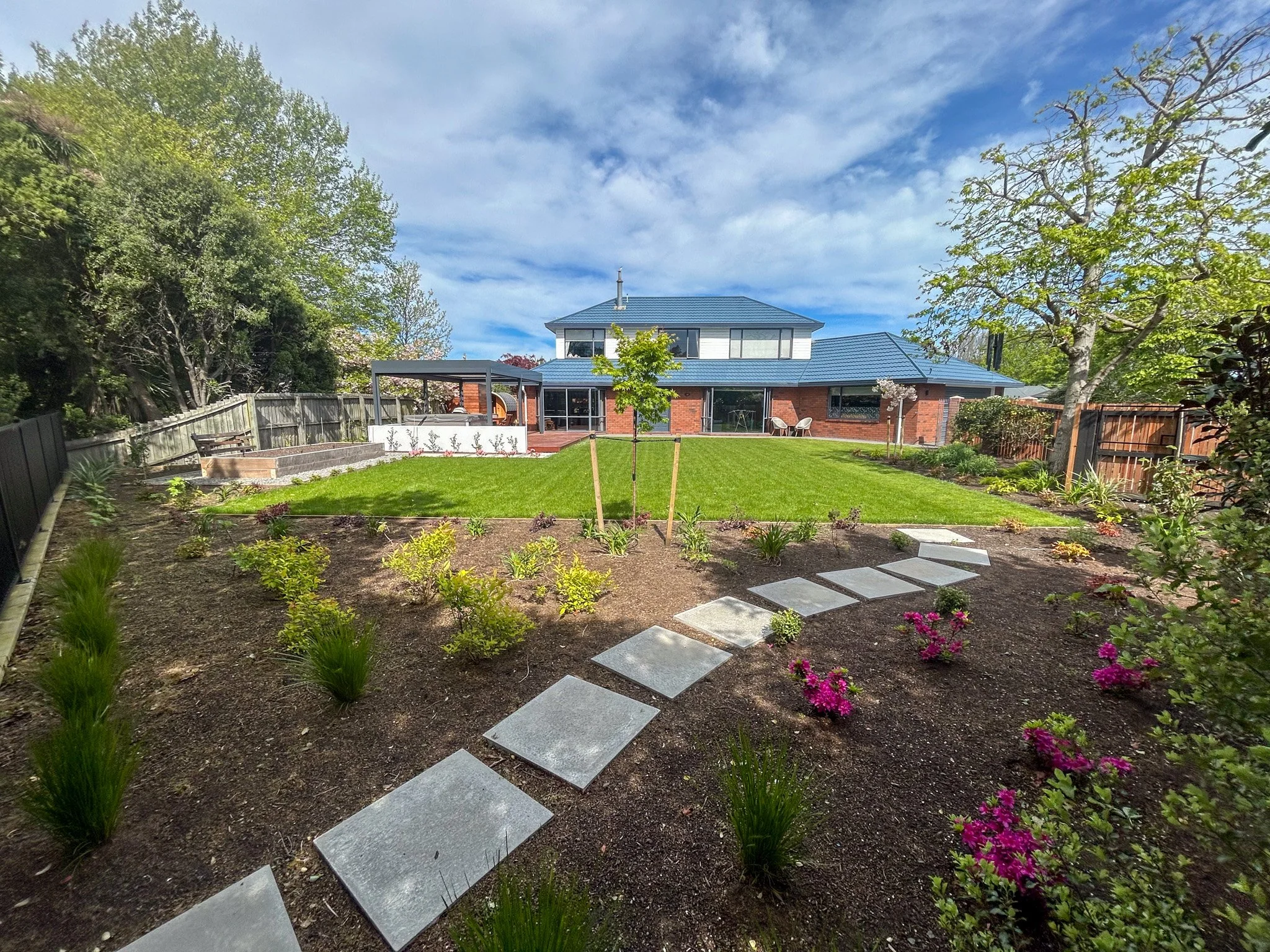 View of a backyard with a grassy lawn, young tree, flower bed, stepping stones, and a house with a blue roof surrounded by trees.
