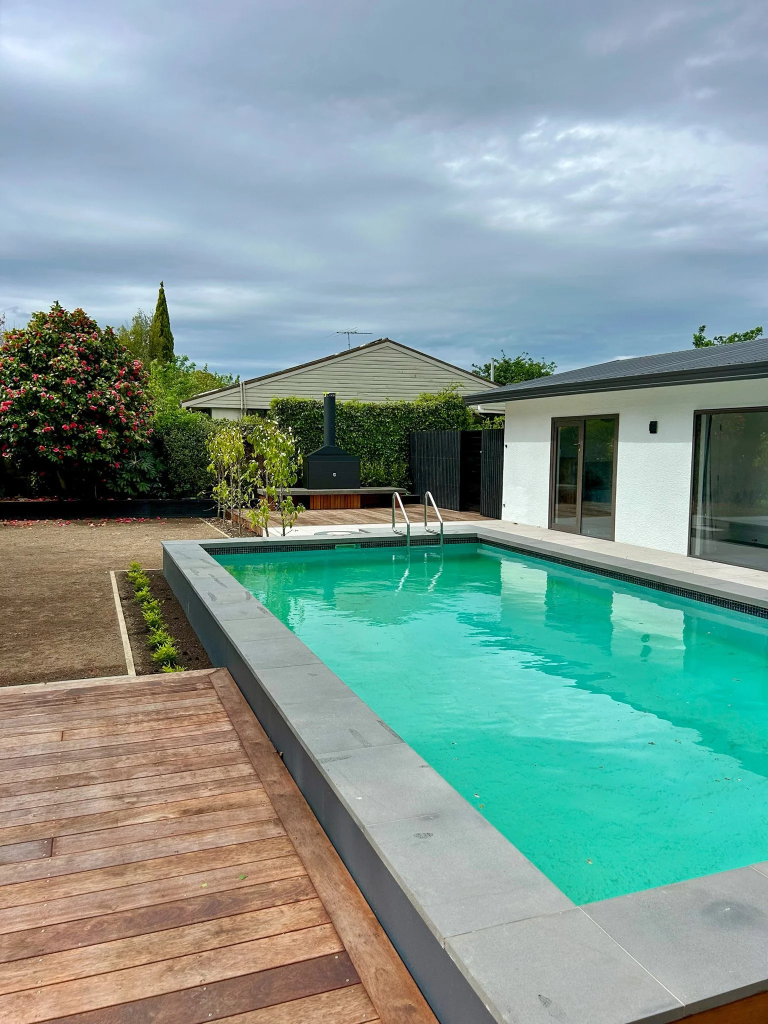 A backyard with a rectangular swimming pool, wooden deck, white house, greenery, and cloudy sky.