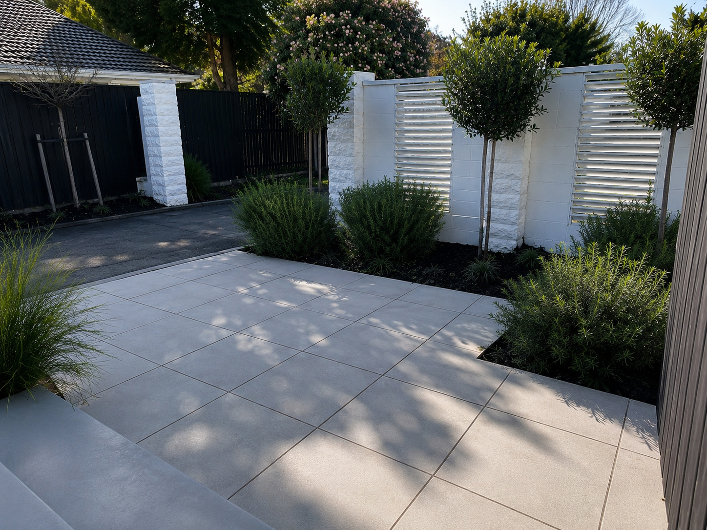 A modern backyard with large beige tiled patio, white stone planter walls, green bushes, and small trees with shadowed areas, enclosed by a black and white fence, during daytime.