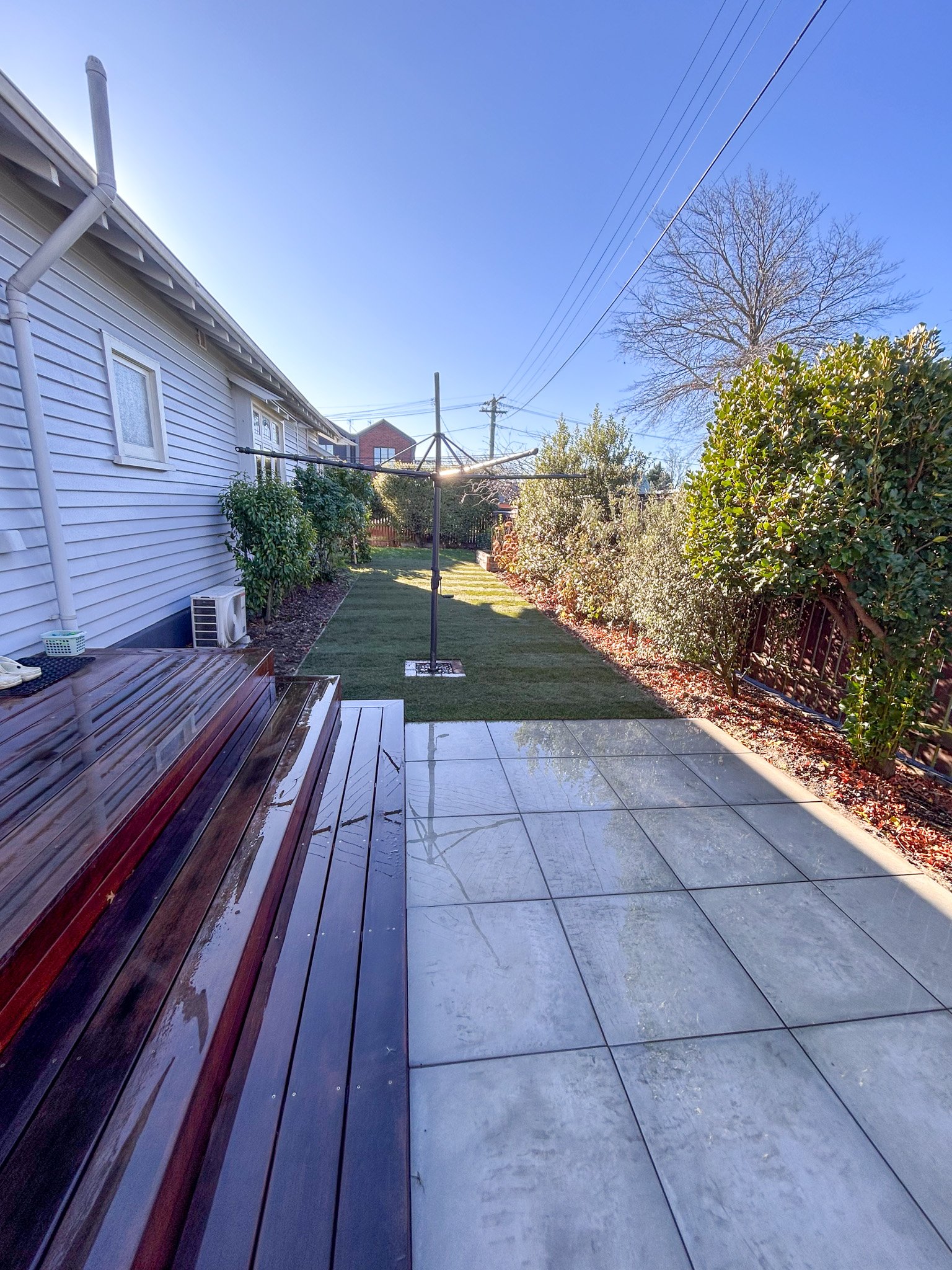 Backyard patio with wooden and tile flooring, a rotary clothesline, bushes along the fence, a house wall on the left, and a clear blue sky with a leafless tree and power lines.