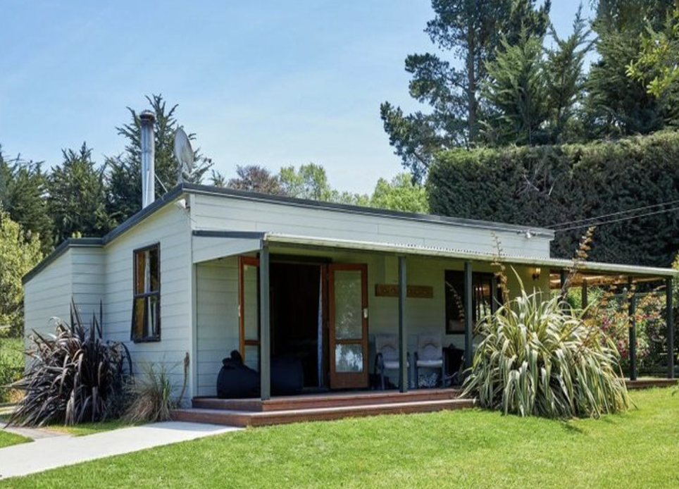 A small, white, single-story house with a porch, surrounded by green grass and tall trees in the background.