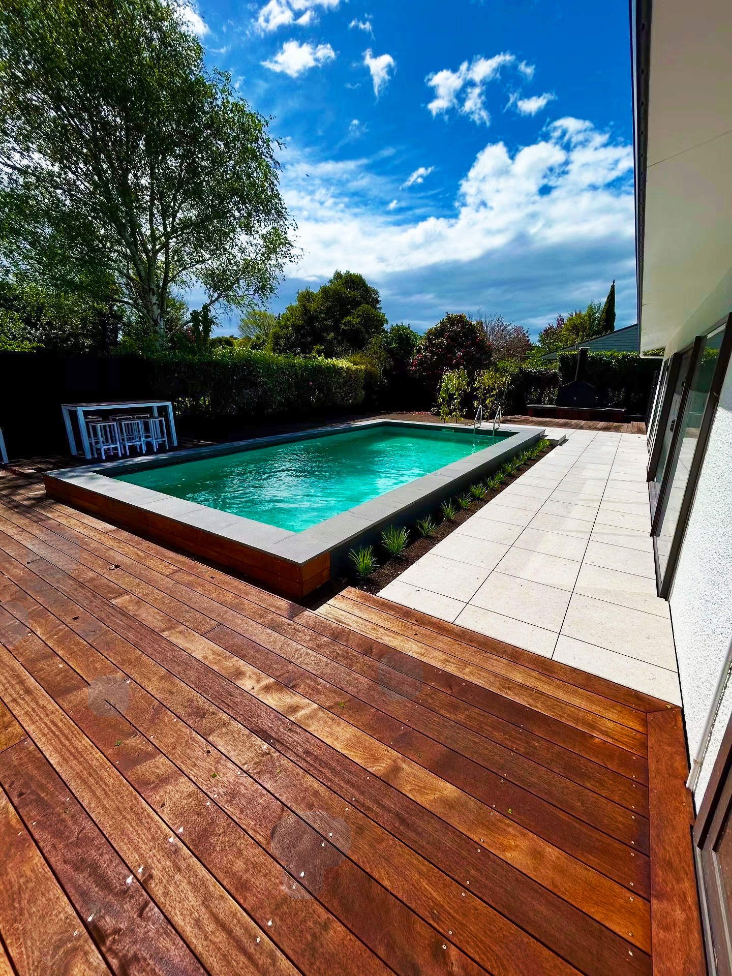 Backyard with a rectangular swimming pool, wooden deck, potted plants, and trees under a partly cloudy blue sky.