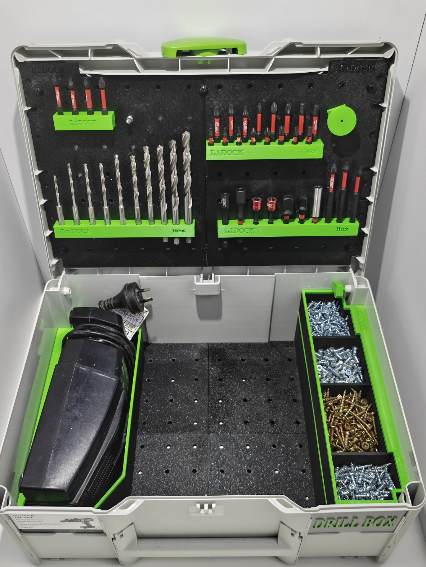 Open plastic drill storage box containing drill bits, screws, and a power drill. The box has green compartments labeled 'L-DOCK' and organized sections for tools and hardware.