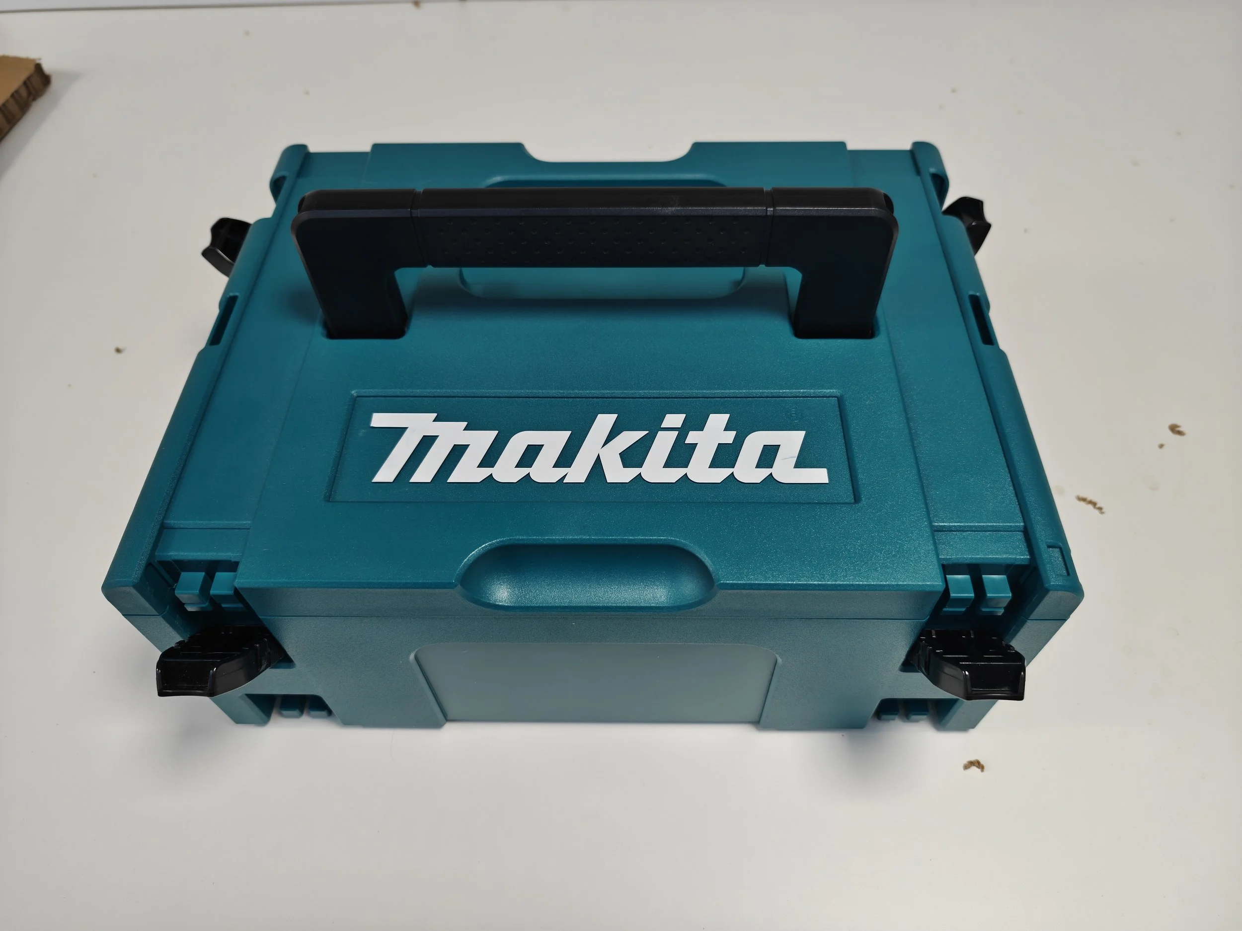 A teal Makita tool box with a black handle on top, placed on a white surface with some small brown debris nearby.