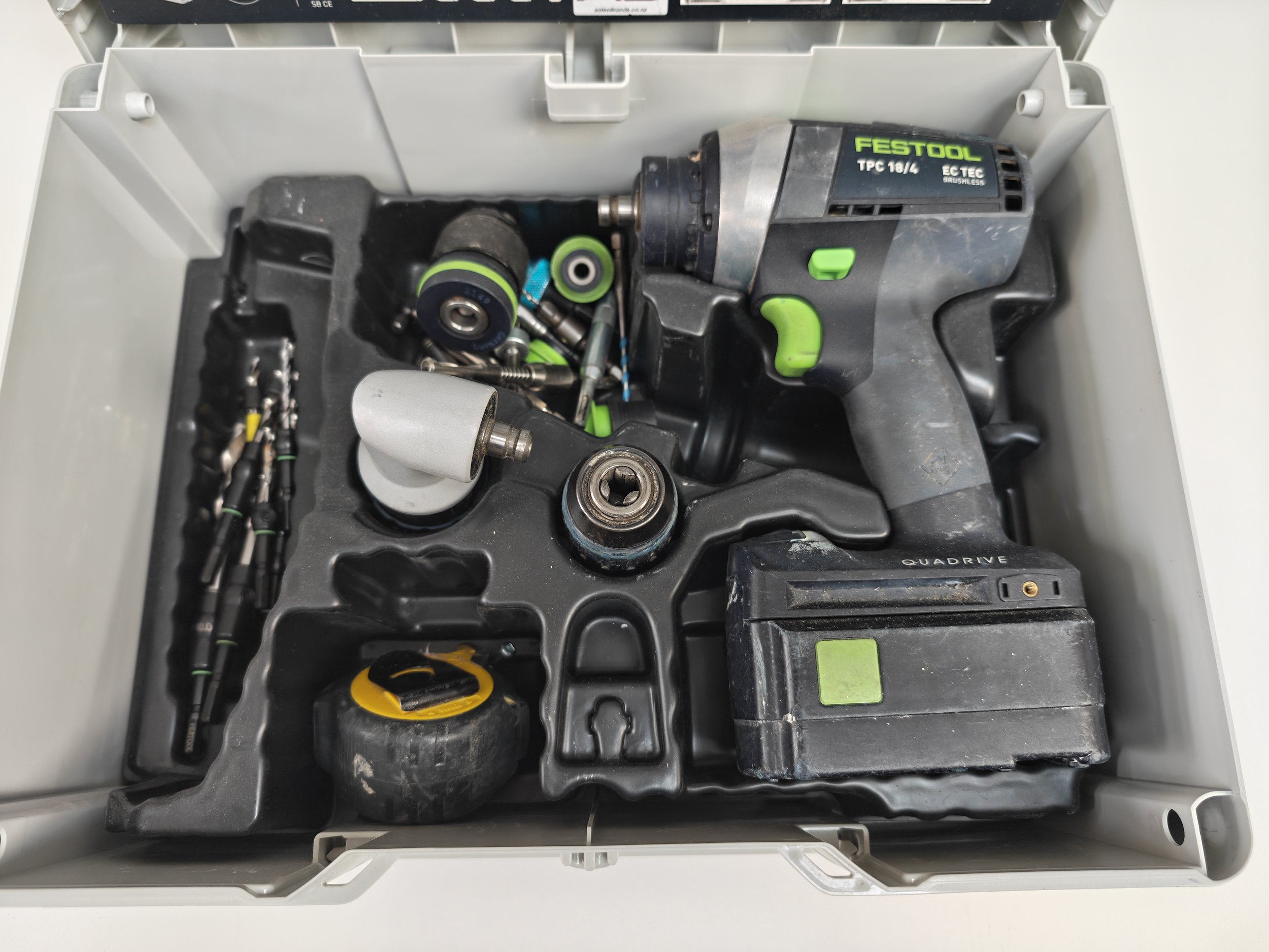 An open gray toolbox containing various tools, including a cordless Festool drill, drill bits, screws, a white rubber roller, and a yellow and black measuring tape.
