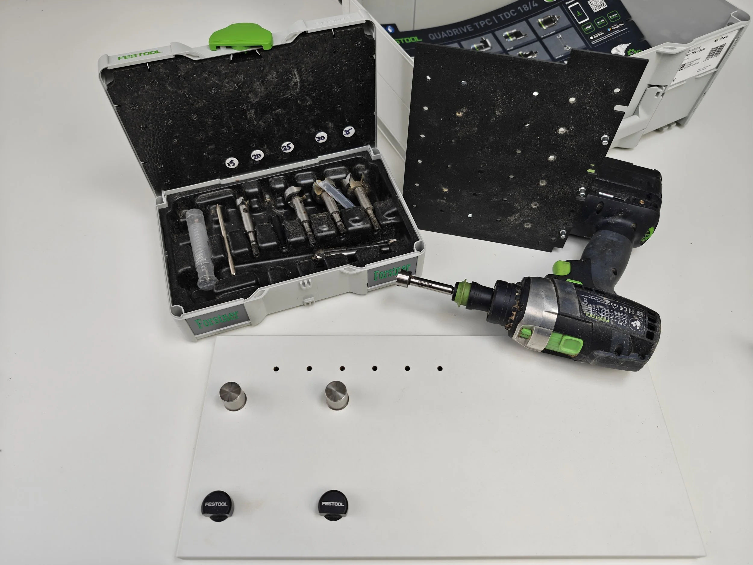 Open black case containing metal drill bits with labels 5, 20, 25, 30, 35, and a syringe inside a foam compartment, a cordless drill with a metal bit attached, a black box, and two black caps labeled 'Festool' on a white surface.