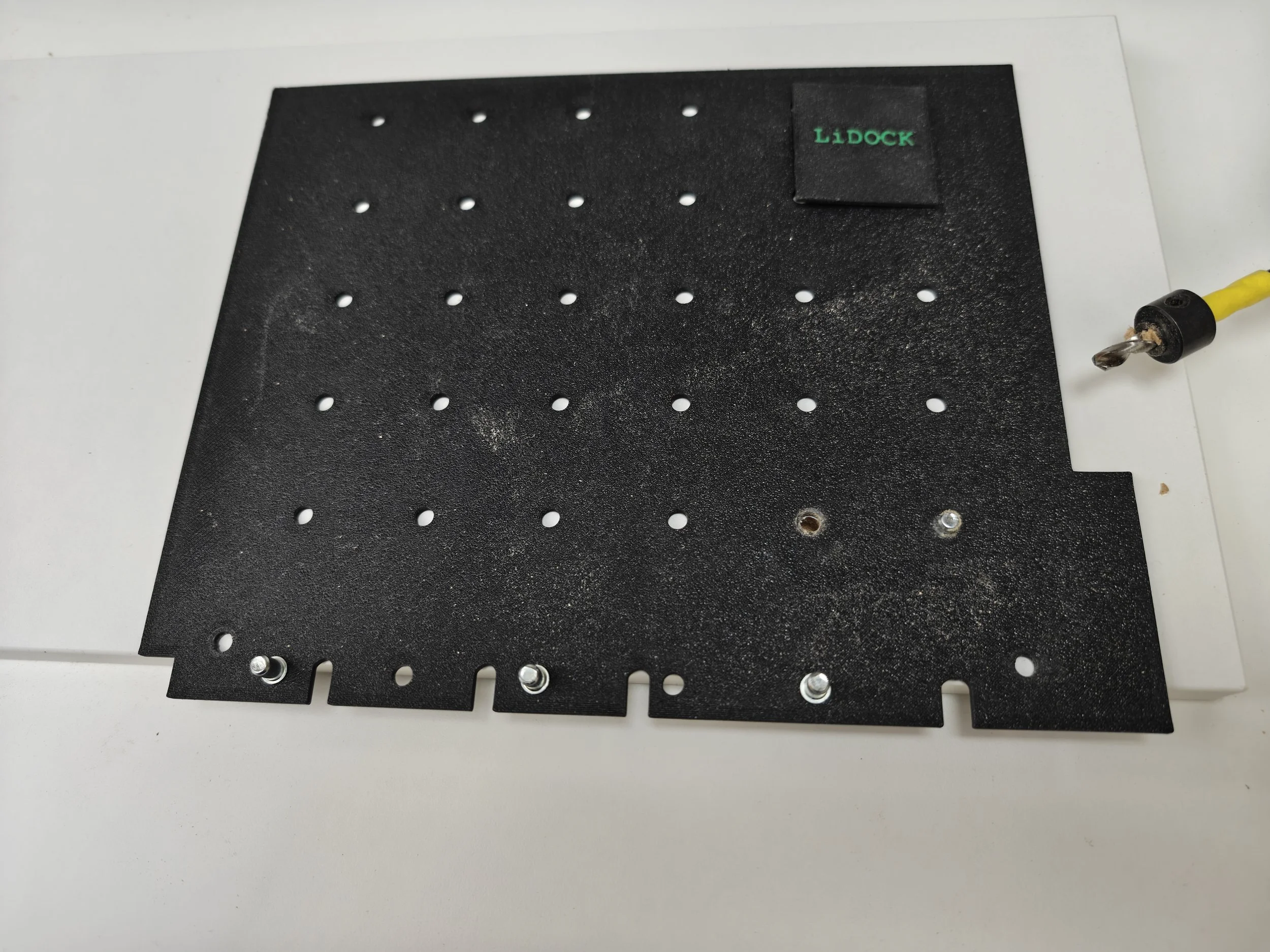 Black perforated board with multiple holes, labeled 'LIDOCK' in green text on a small black patch, and a yellow cable with a metal connector on the right side.