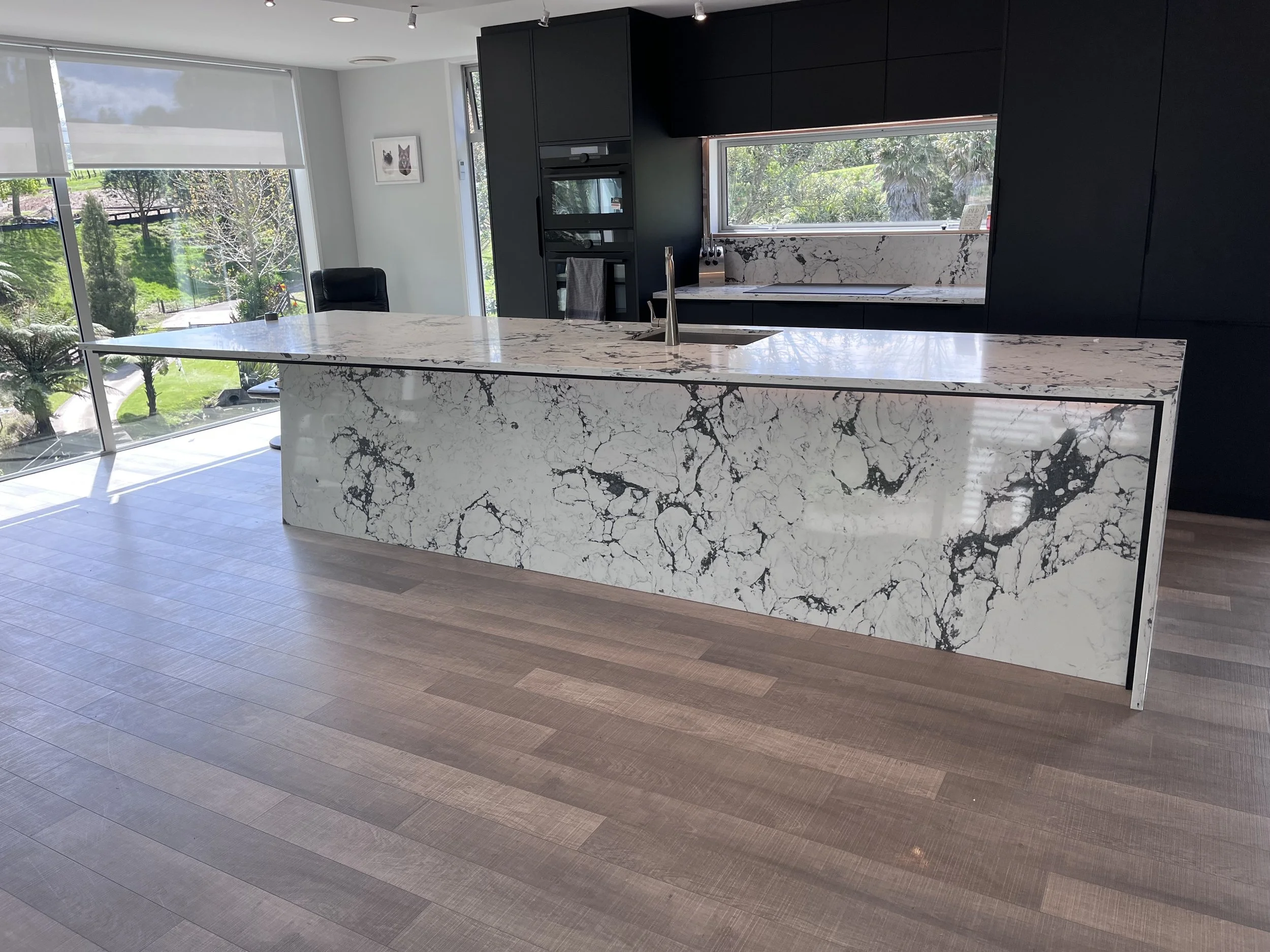 Modern kitchen with marble island and black cabinets, large window showing greenery outside, and a sliding glass door with a view of a park or garden.
