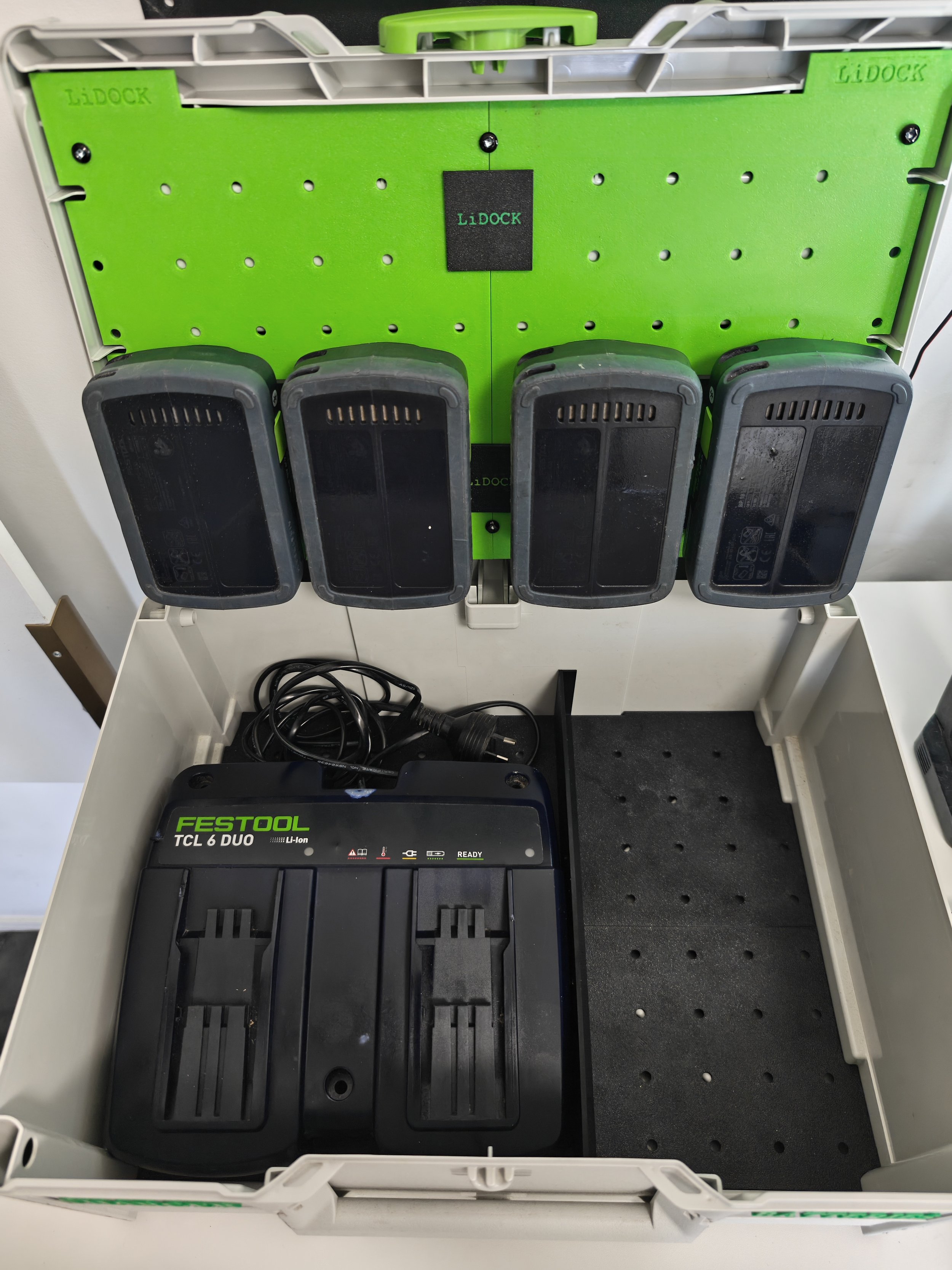 Open white plastic electrical box containing a black Festool TCL 6 DUO lithium-ion battery charger, with a green and black lidock panel behind it and four black battery slots.