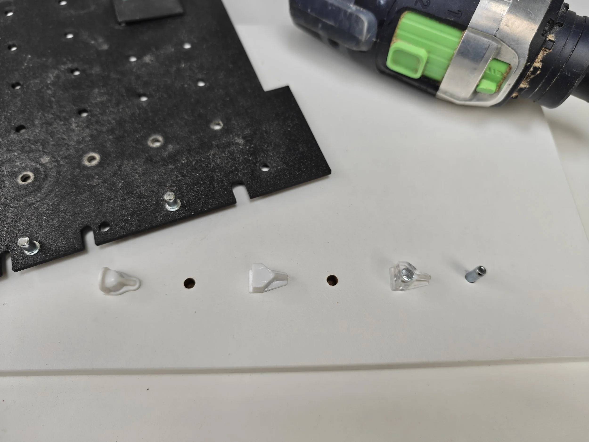 Close-up of a black perforated metal sheet and a cordless drill with a green drill bit, on a white surface with small miscellaneous plastic and metal parts.