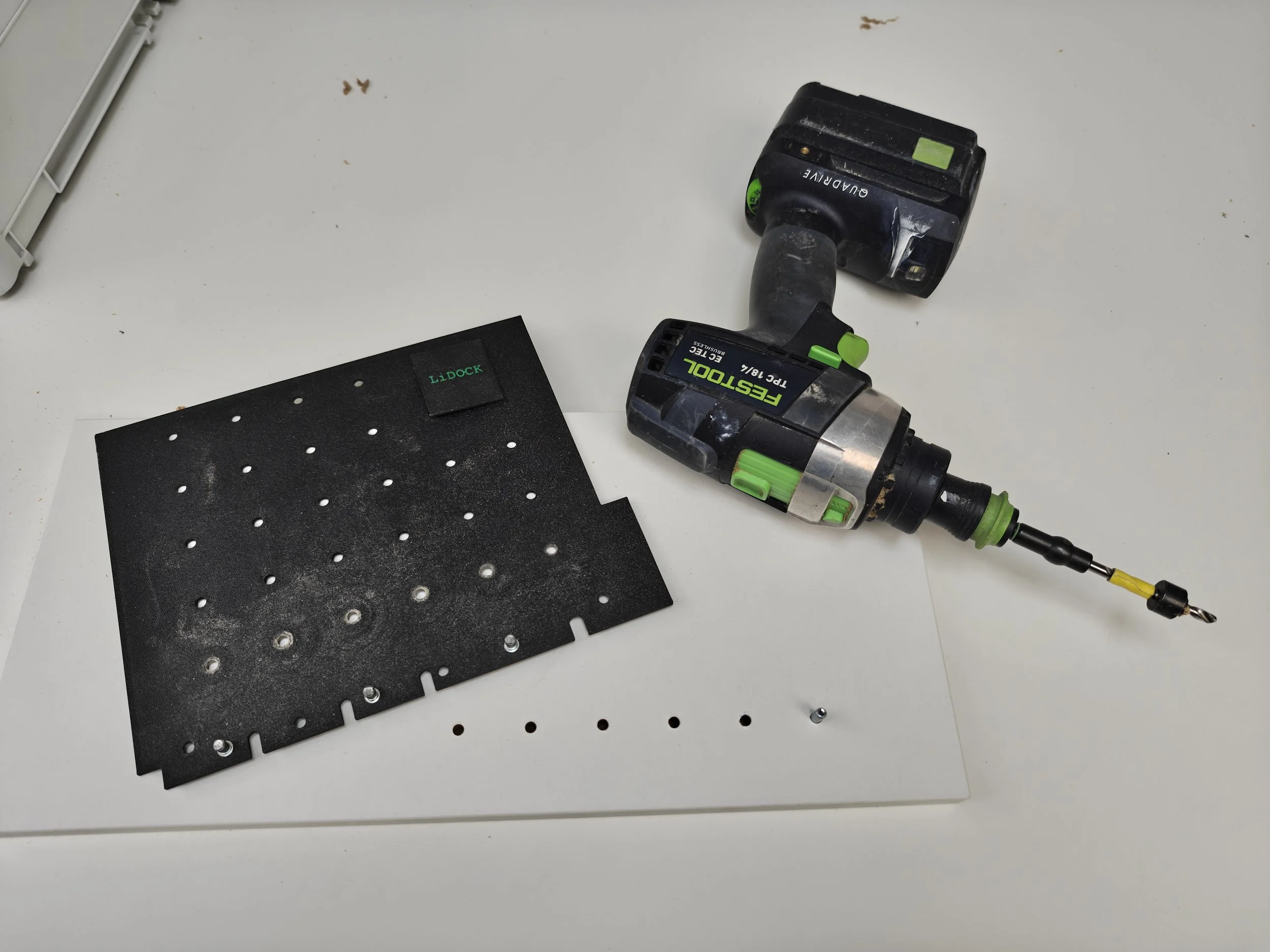 A cordless power drill lying on a white table next to a black perforated sheet of material and a small screw.