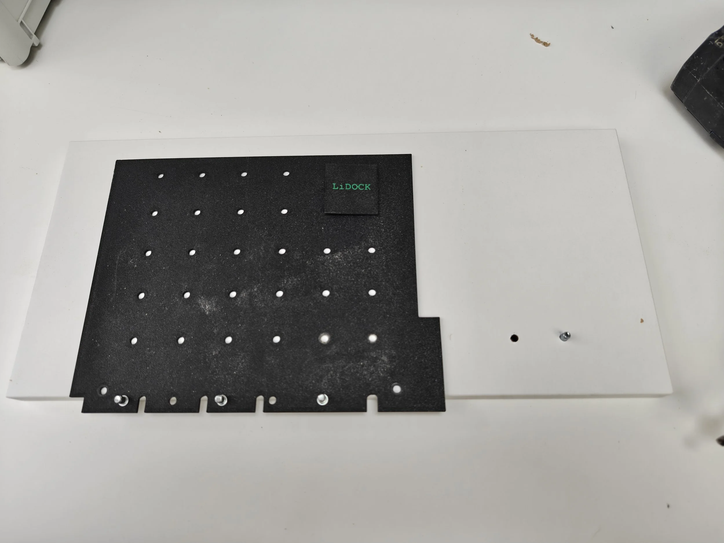 A black Perforated metal plate with a small green label reading 'LiDOCK' on a white surface.