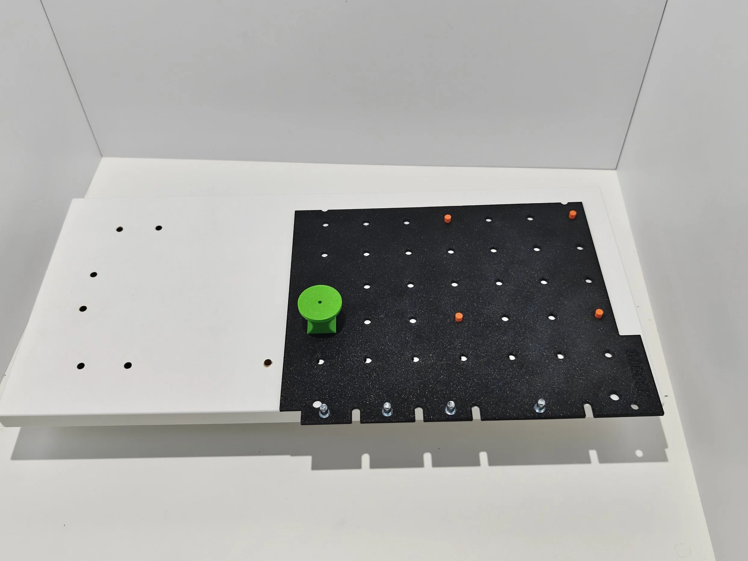 A white table or surface with a black pegboard placed on top, containing small orange and silver pegs and a green circular piece.
