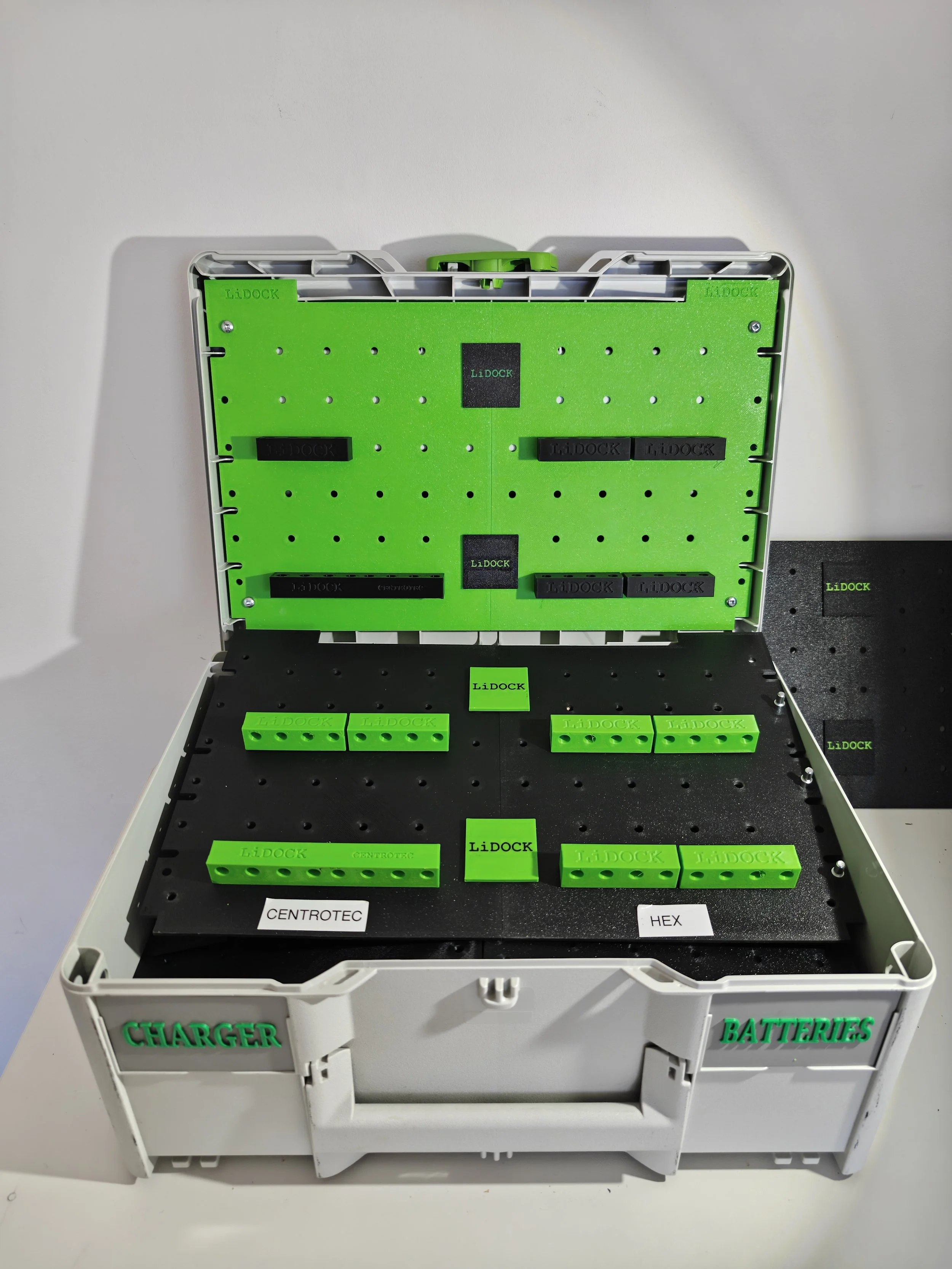An open white plastic container with green labeled parts for assembling medical or electronic equipment. The container has sections labeled 'CHARGER' and 'BATTERIES' and contains green components labeled 'LiDOCK' used for modular assembly.
