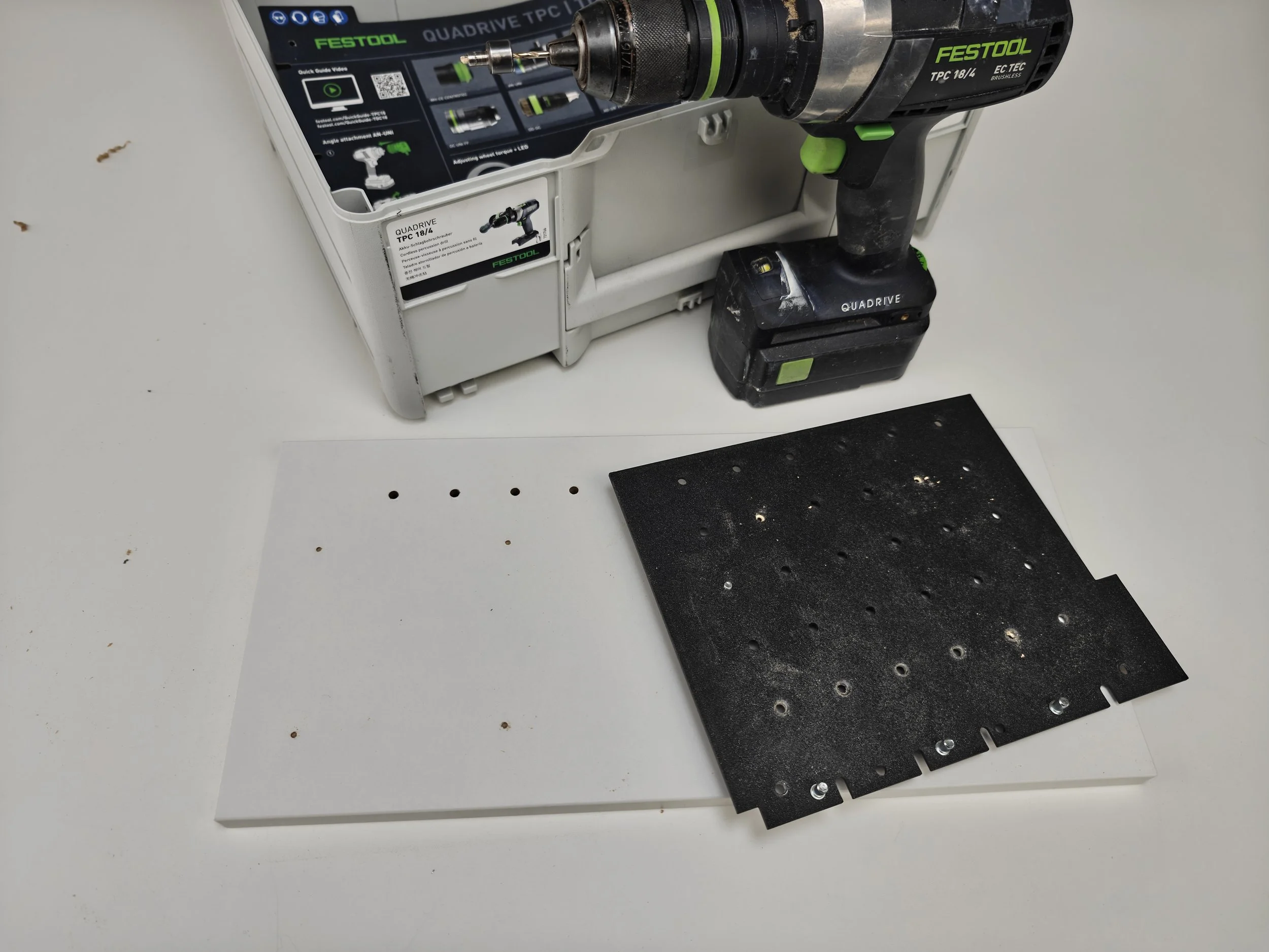 A black and green Festool drill is placed on a white surface, with the drill's bit pointed downward onto a piece of black material with evenly spaced holes. The drill is positioned to the right of a white plastic case with a detailed label and product information.