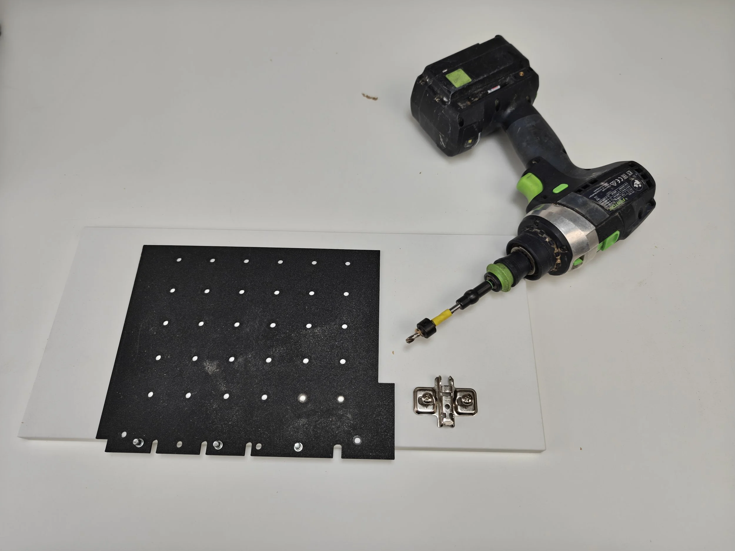 Power drill, black metal perforated board, and hinge on a white surface.