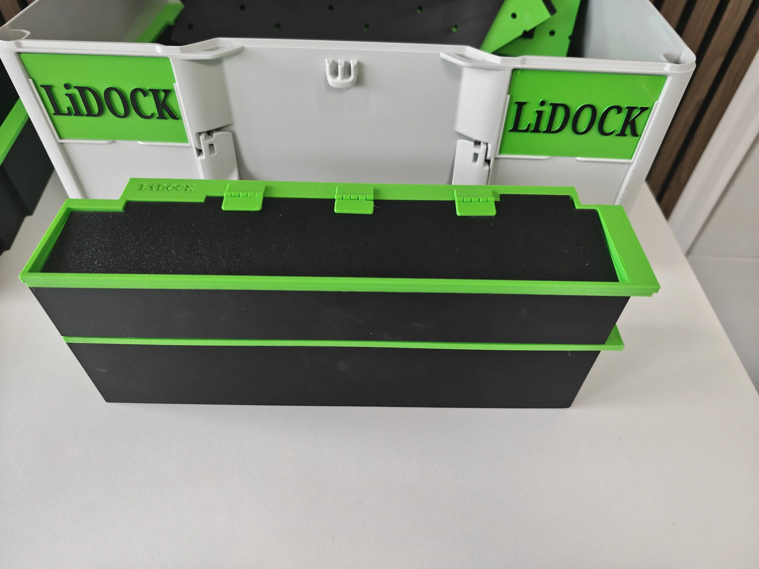A white and green toolbox with the word "LIDOCK" on the side, open to show a black foam interior, sitting on a white surface.