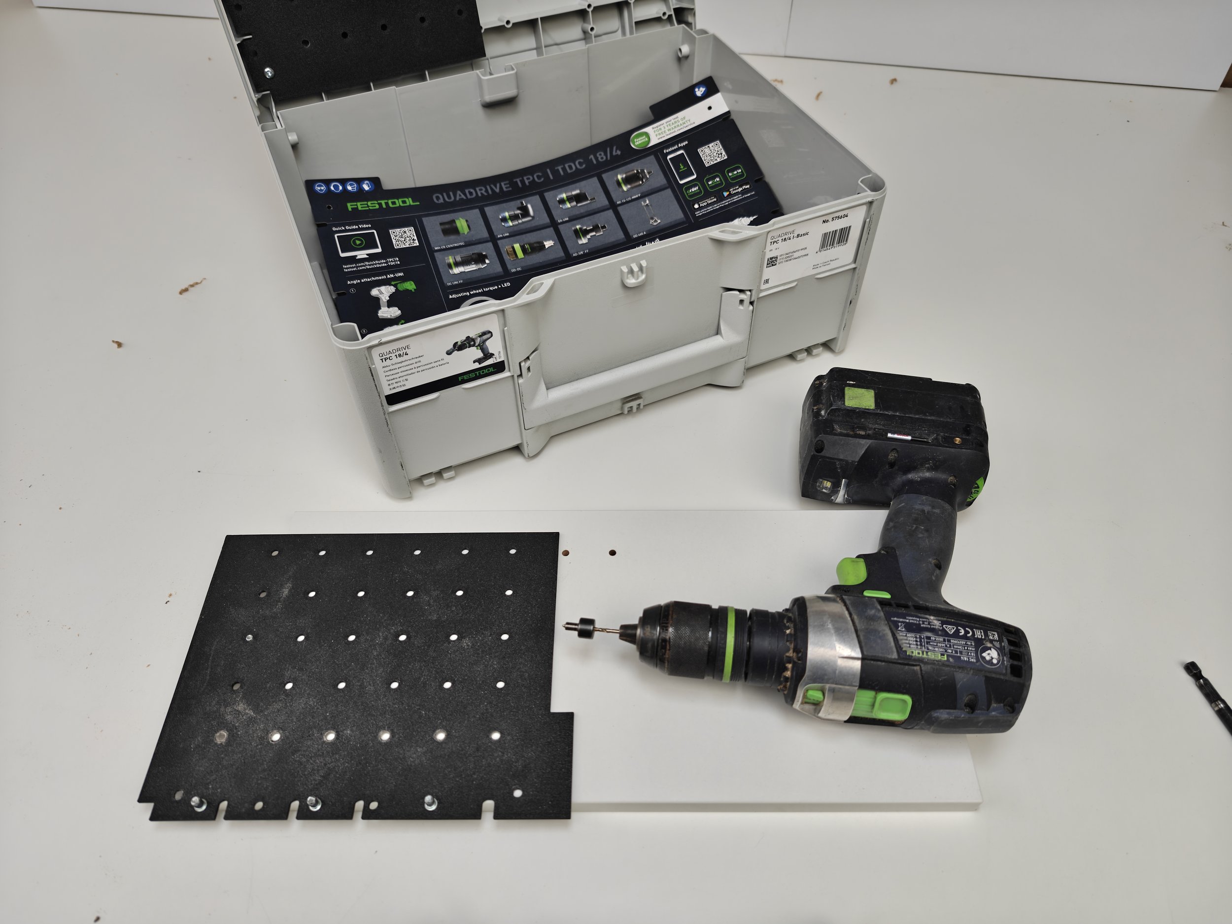 Open plastic toolbox with instruction sheet inside, black perforated mat, cordless drill, and driver bit on a white work surface.