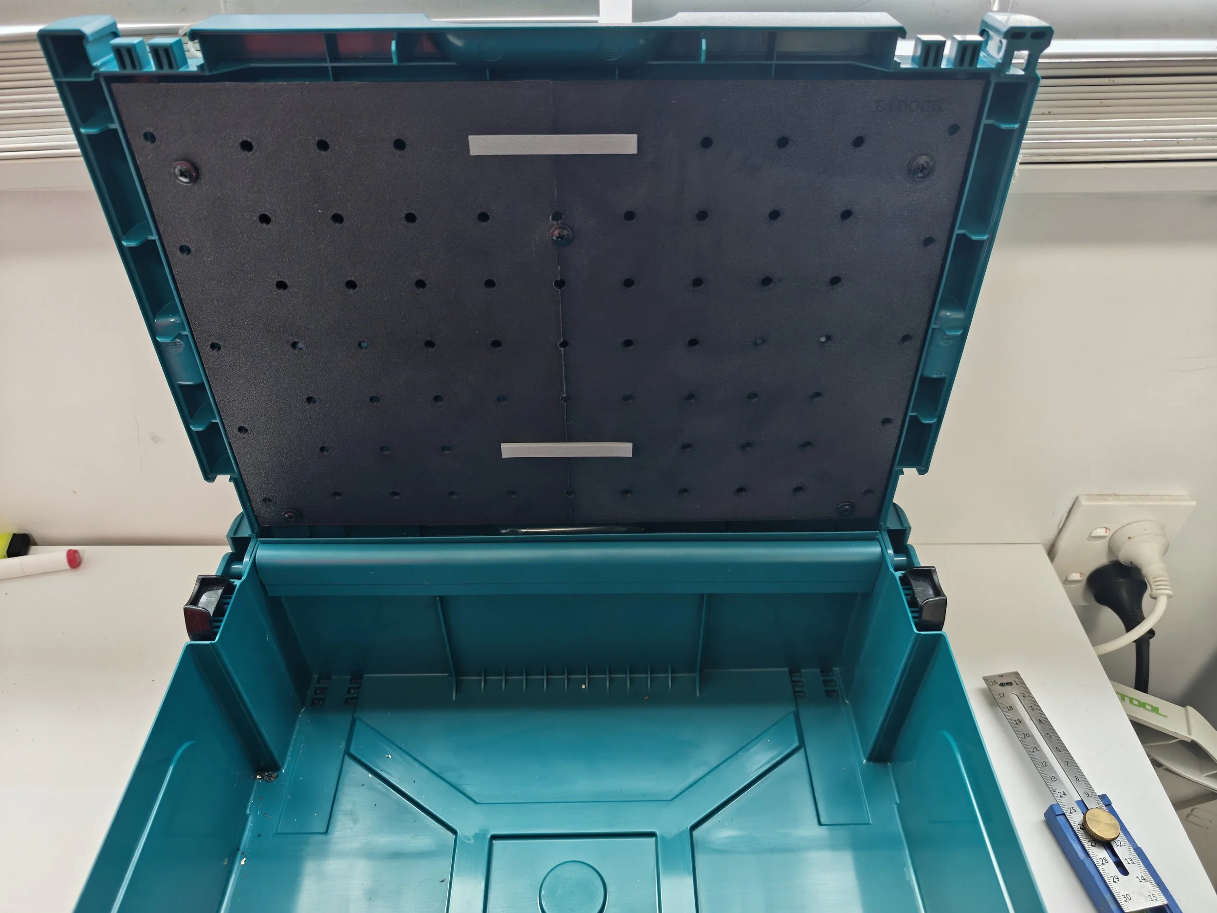 A teal plastic toolbox with its lid open, showing an empty interior, placed on a white surface with a ruler, wall socket, and some cables nearby.